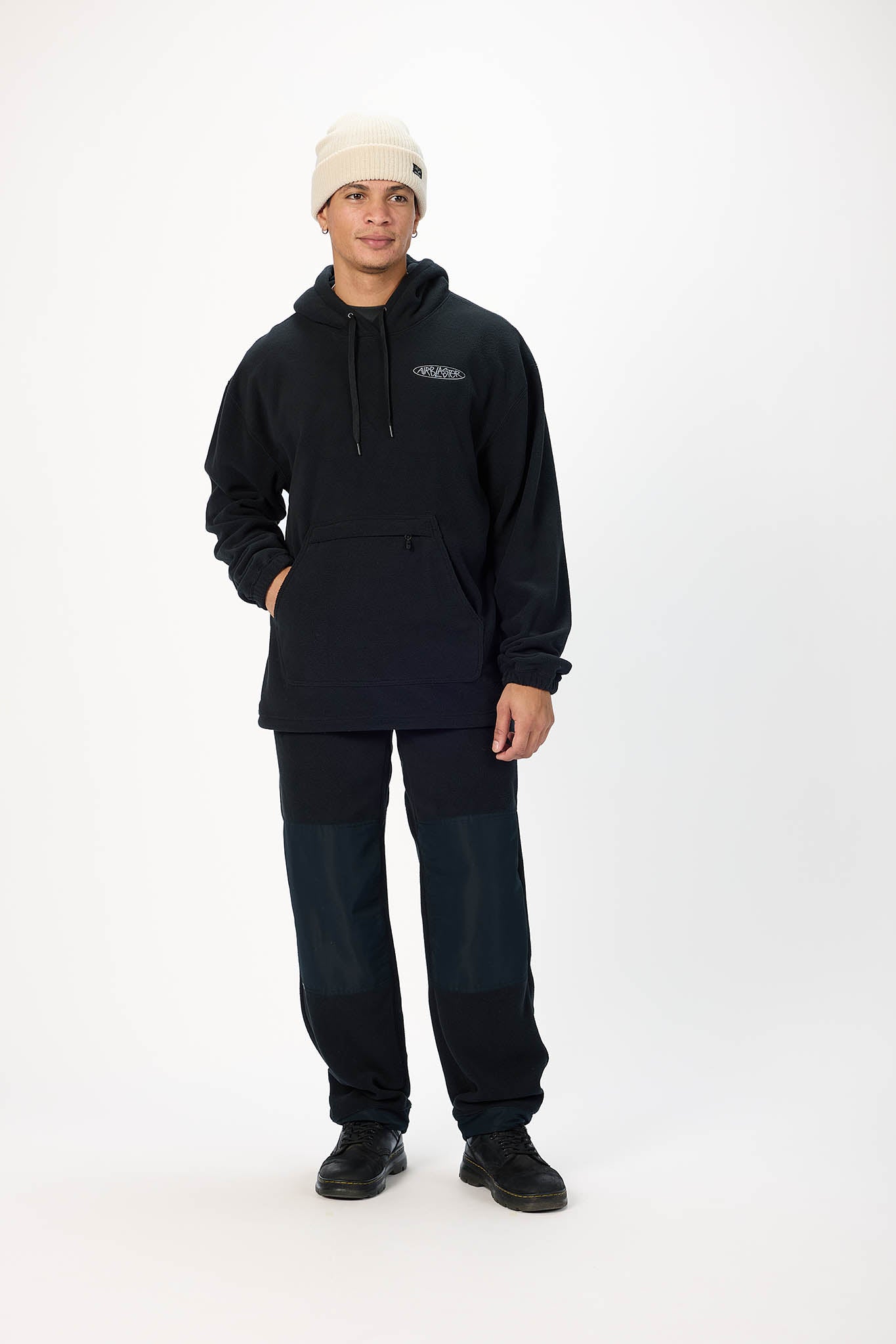 A man modeling a dark navy pullover hoodie with a small white chest logo, matching dark navy pants, black shoes, and a cream-colored beanie against a plain white studio background.