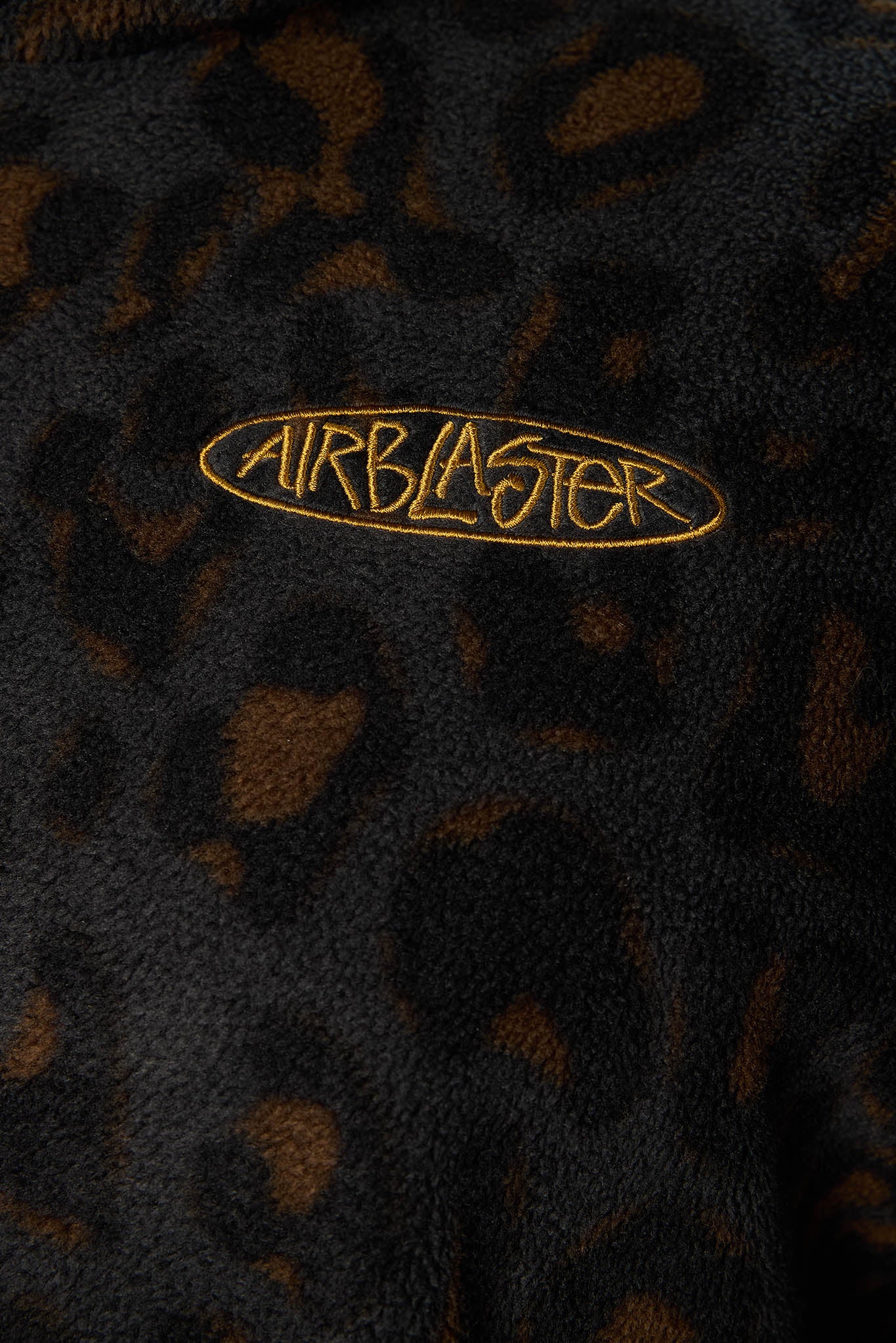Close-up of a black and brown leopard print fleece garment with a gold embroidered oval logo reading AIRBLASTER on the chest.