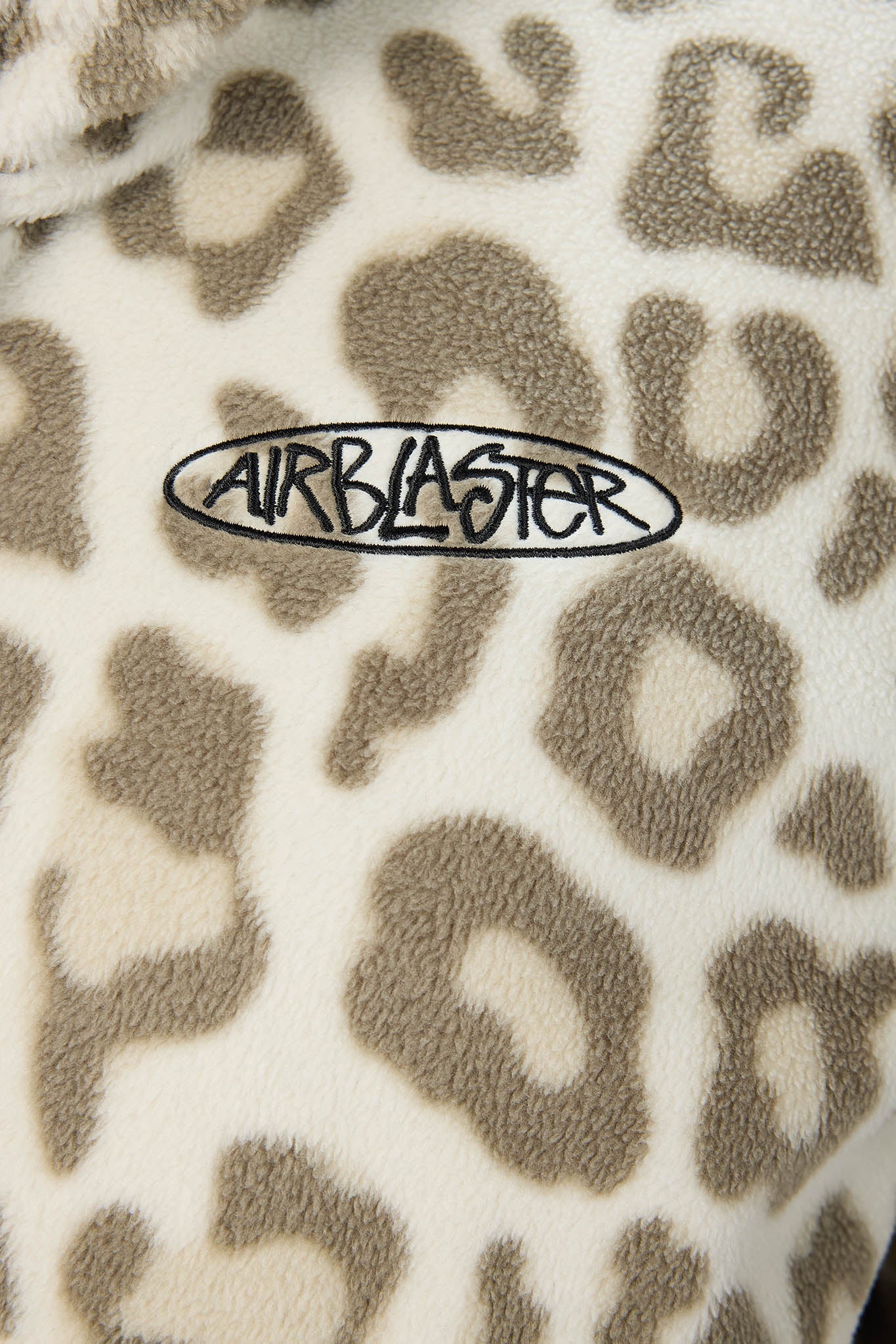Close-up of a plush fleece fabric with a beige and brown leopard print pattern and a black embroidered oval logo reading AIRBLASTER.