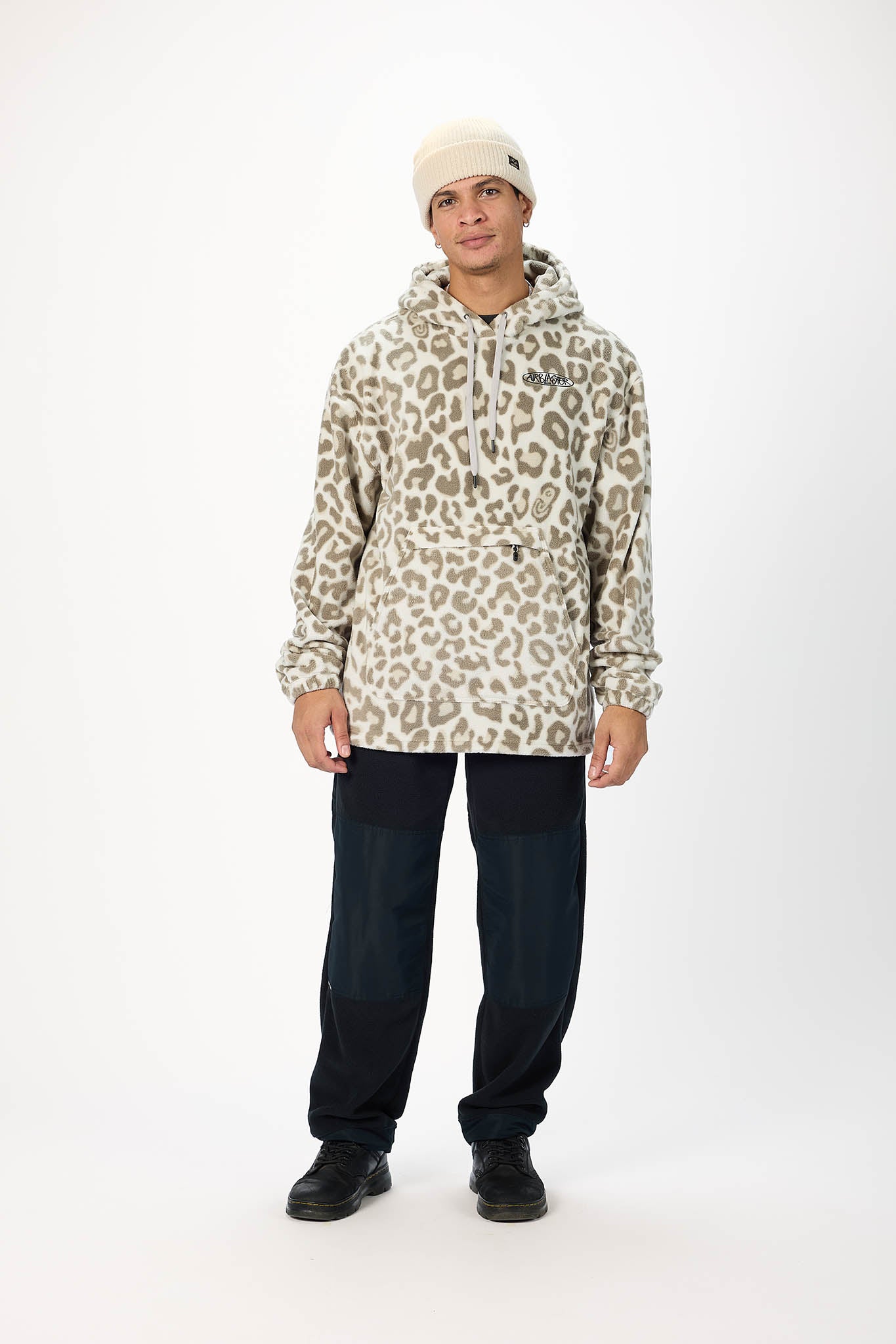 A man modeling a beige and brown leopard-print hooded jacket, paired with dark navy pants, black shoes, and a cream beanie, standing against a plain white studio background.