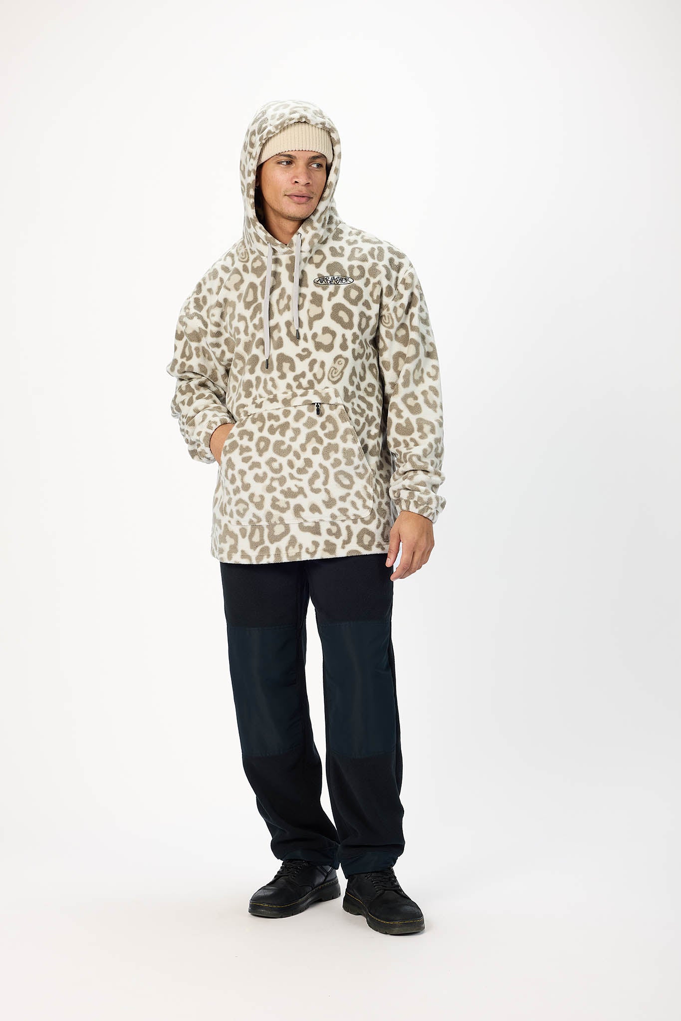 A man modeling a hooded jacket with a beige and brown leopard print pattern, paired with dark pants and black shoes, against a plain white studio background.