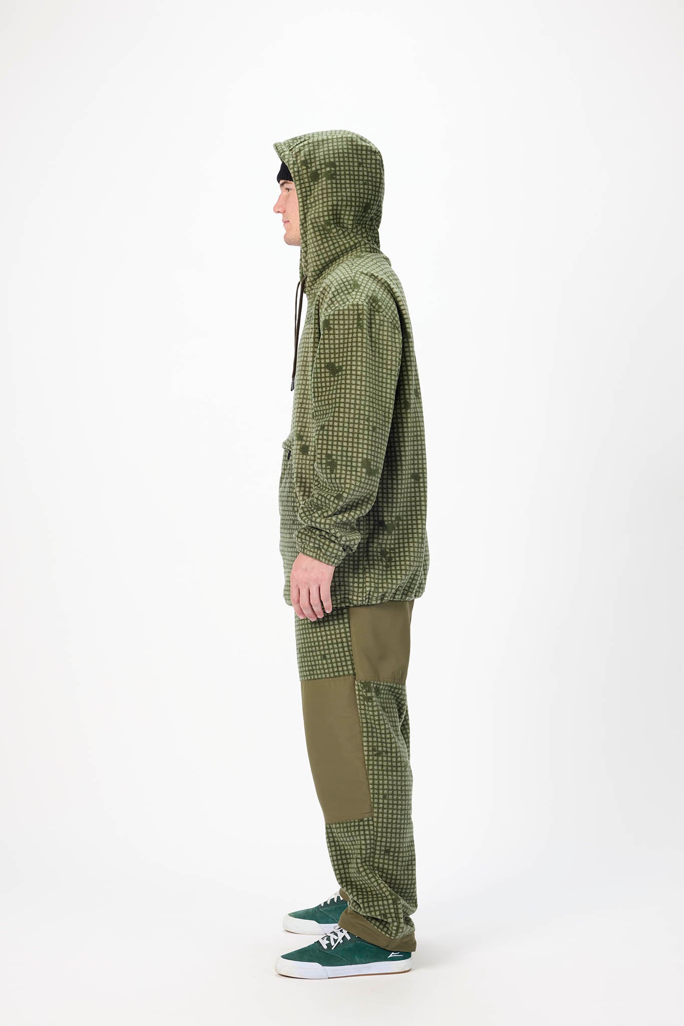 A person shown in side profile wearing an olive green hooded jacket with a subtle tonal pattern, matching olive pants, and green sneakers against a plain white studio background.