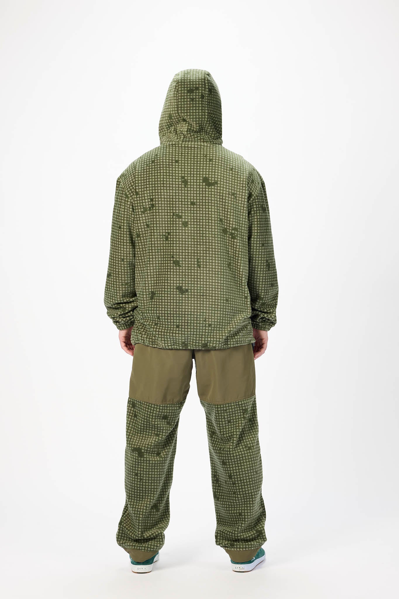 A person shown from the back wearing an olive green hooded jacket and matching green pants with a small check pattern and darker green patch panels, photographed against a plain light gray studio background.
