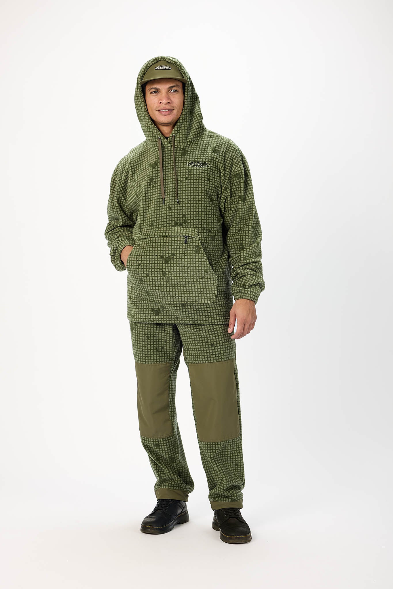 A man modeling an olive green hooded fleece jacket and matching pants with green knee patches, photographed against a plain white studio background.