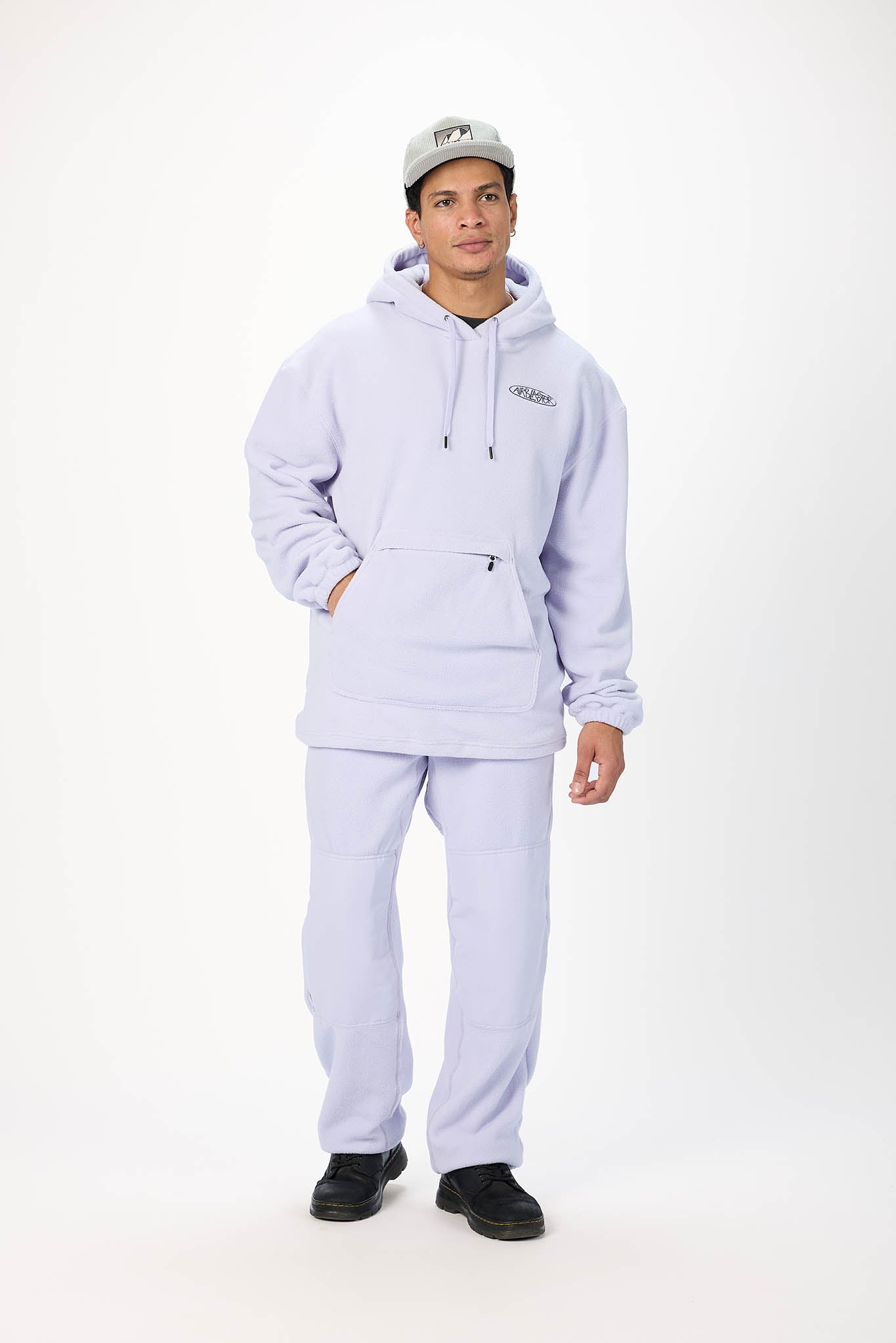A man modeling a light lavender matching hoodie and sweatpants set, paired with a gray baseball cap and black shoes, against a plain white studio background.