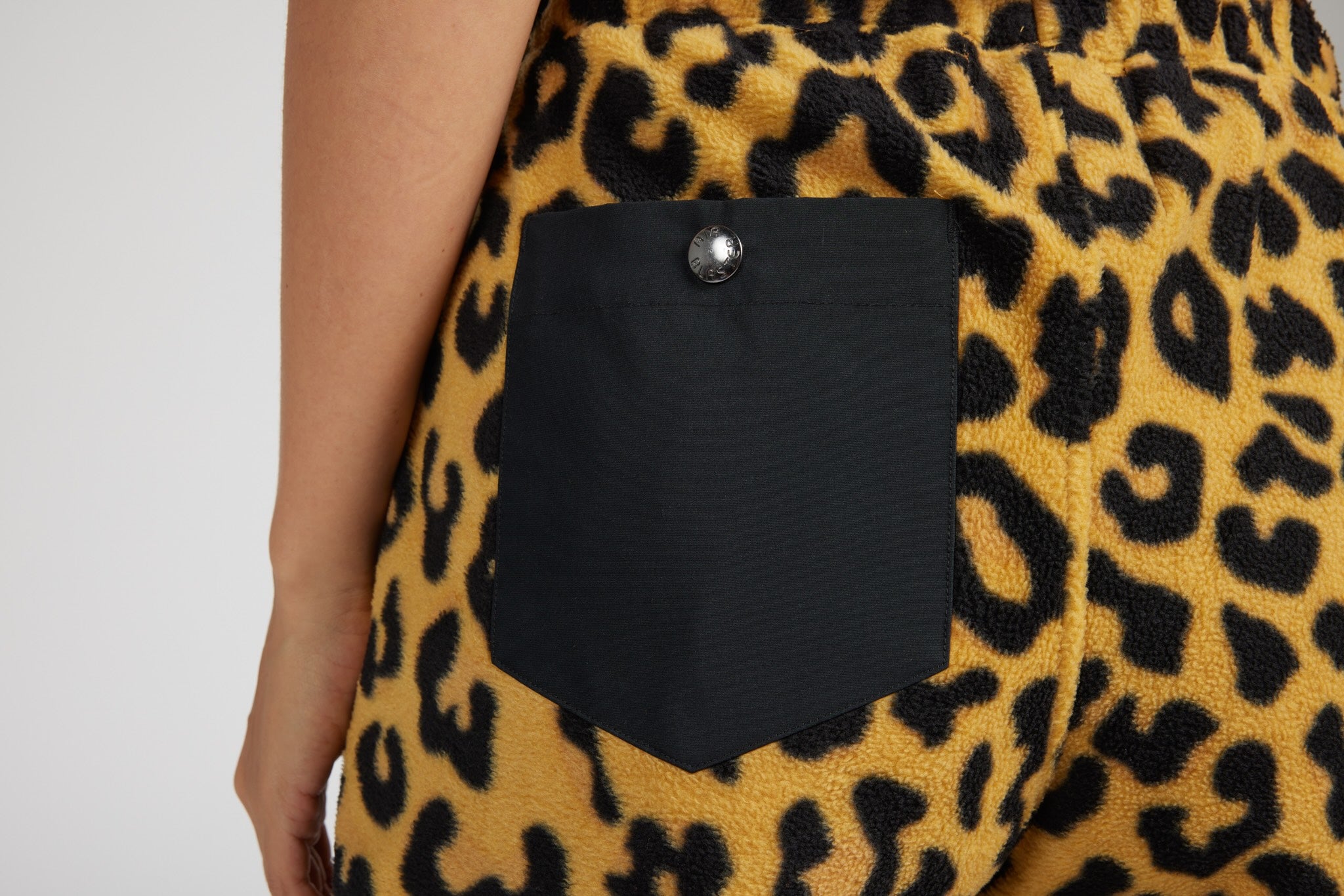 Close-up of a leopard-print fleece garment with a black buttoned patch pocket, worn by a model against a light gray background.