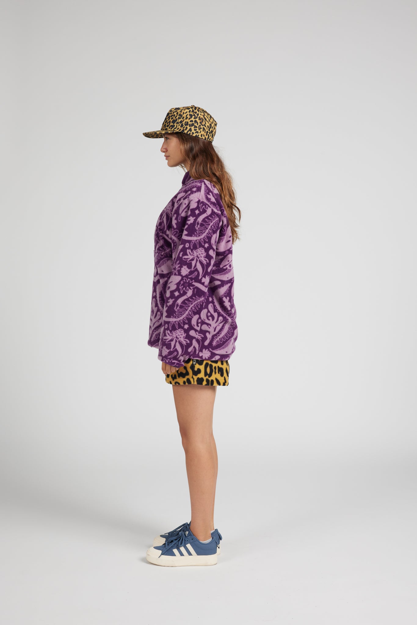 A model is shown in profile wearing a purple paisley fleece jacket, leopard-print shorts, a leopard-print cap, and blue sneakers against a plain light gray studio background.