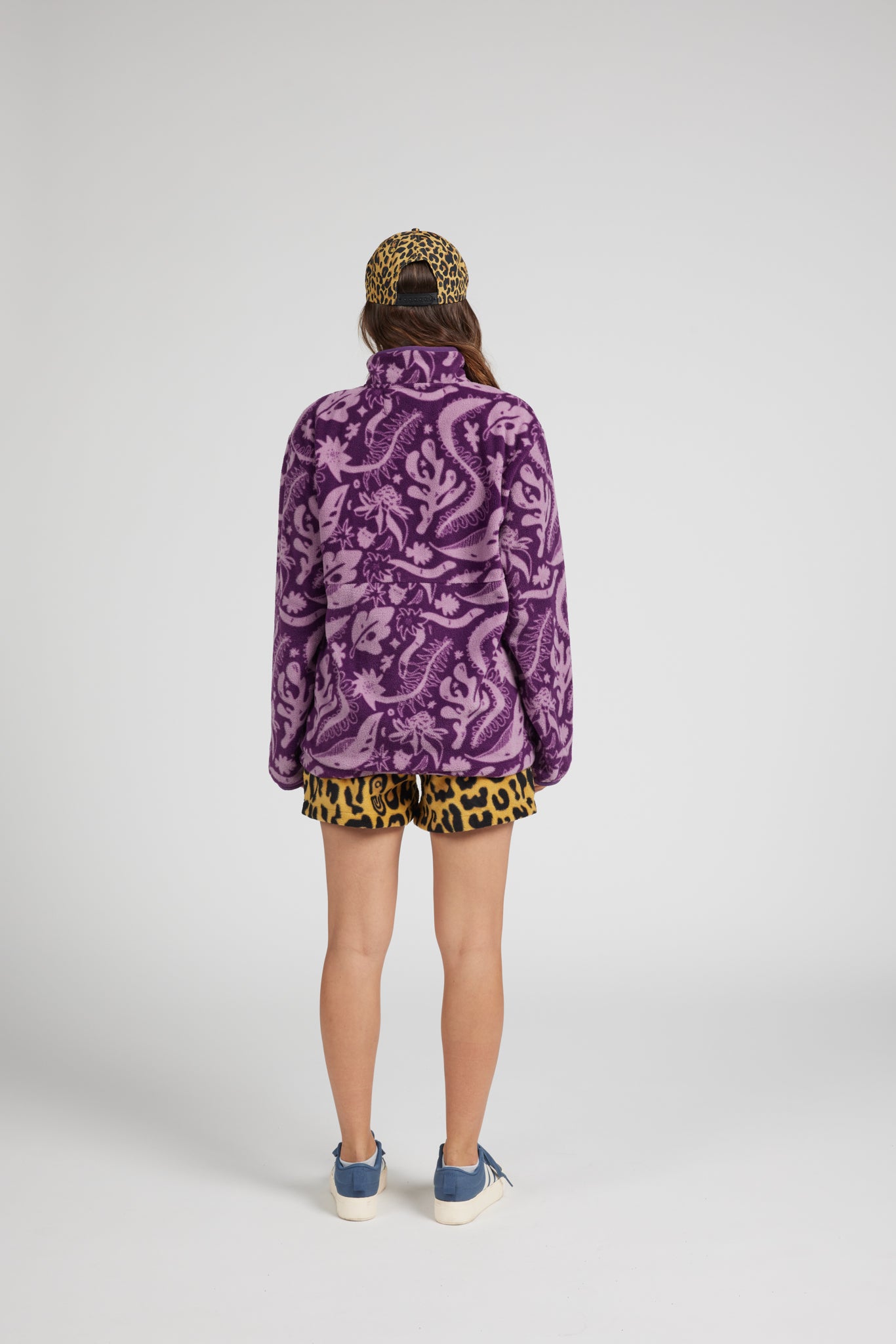 A model shown from the back wearing a purple and black paisley print fleece jacket, matching leopard-print shorts, a leopard-print cap, and blue sneakers against a light gray studio background.