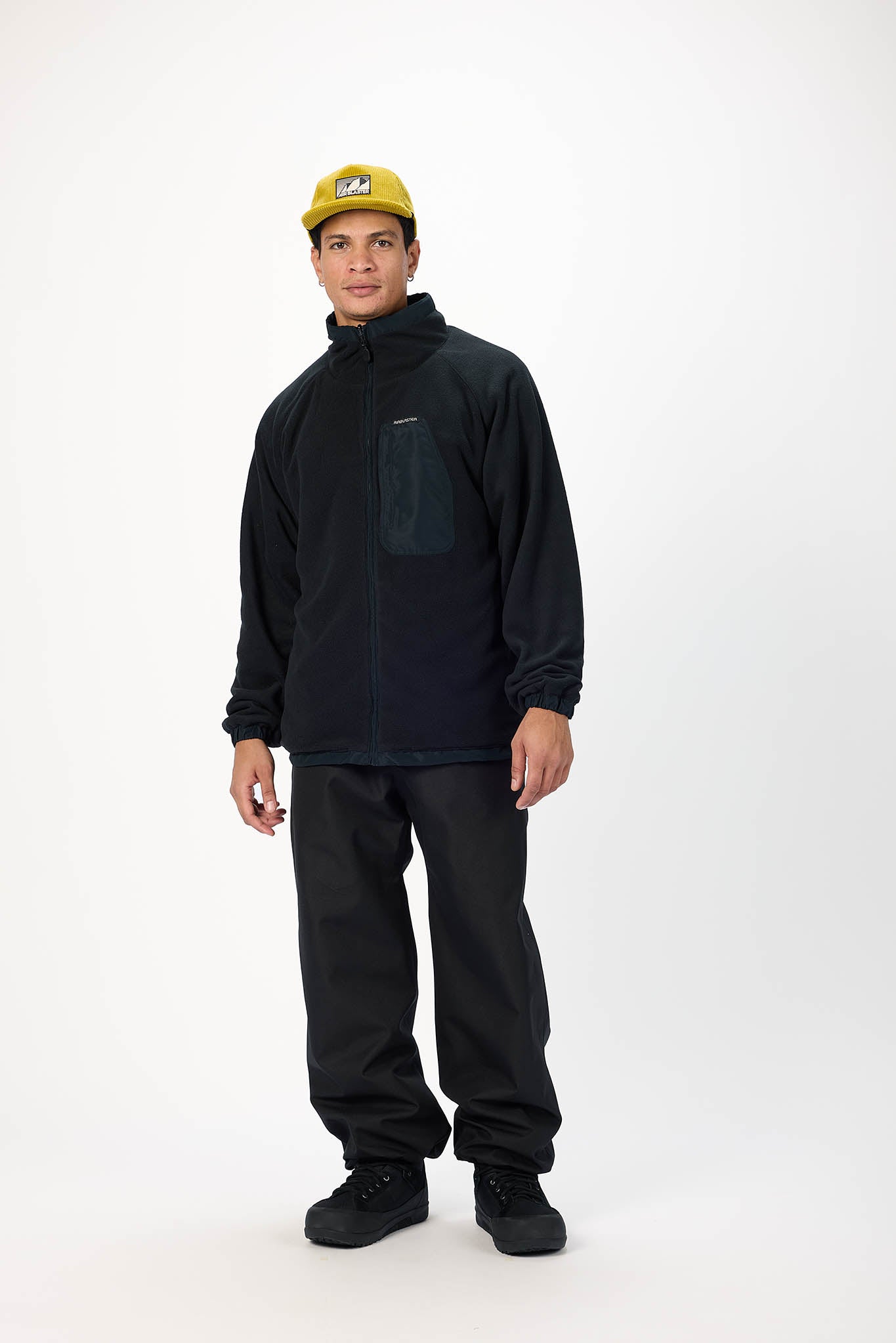 A man modeling a black fleece jacket with a high collar and a dark chest pocket, paired with black pants and a yellow cap, against a plain white studio background.