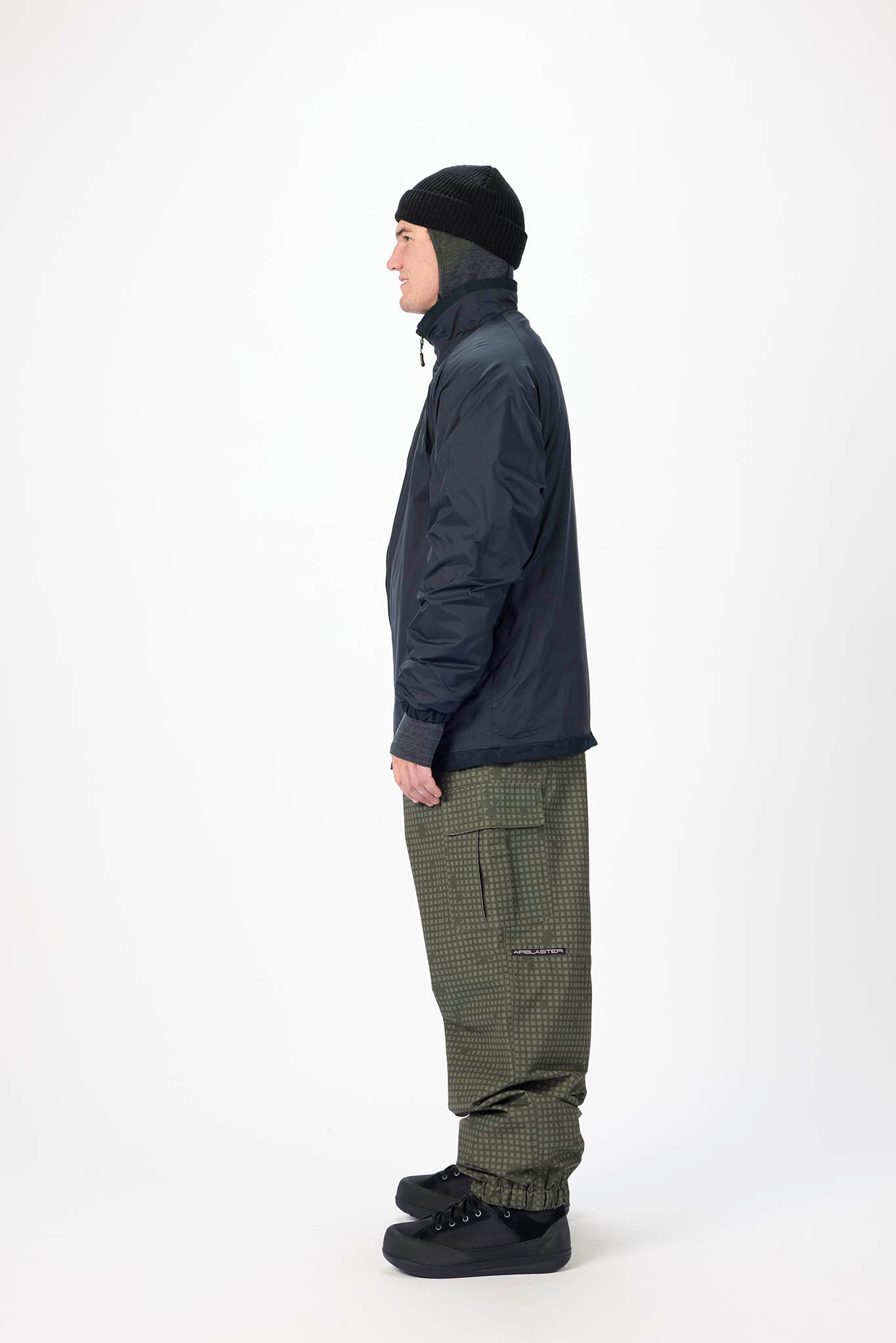 A man shown in profile wearing a dark navy jacket, olive green patterned pants, a black beanie, and black shoes, standing against a plain white studio background.