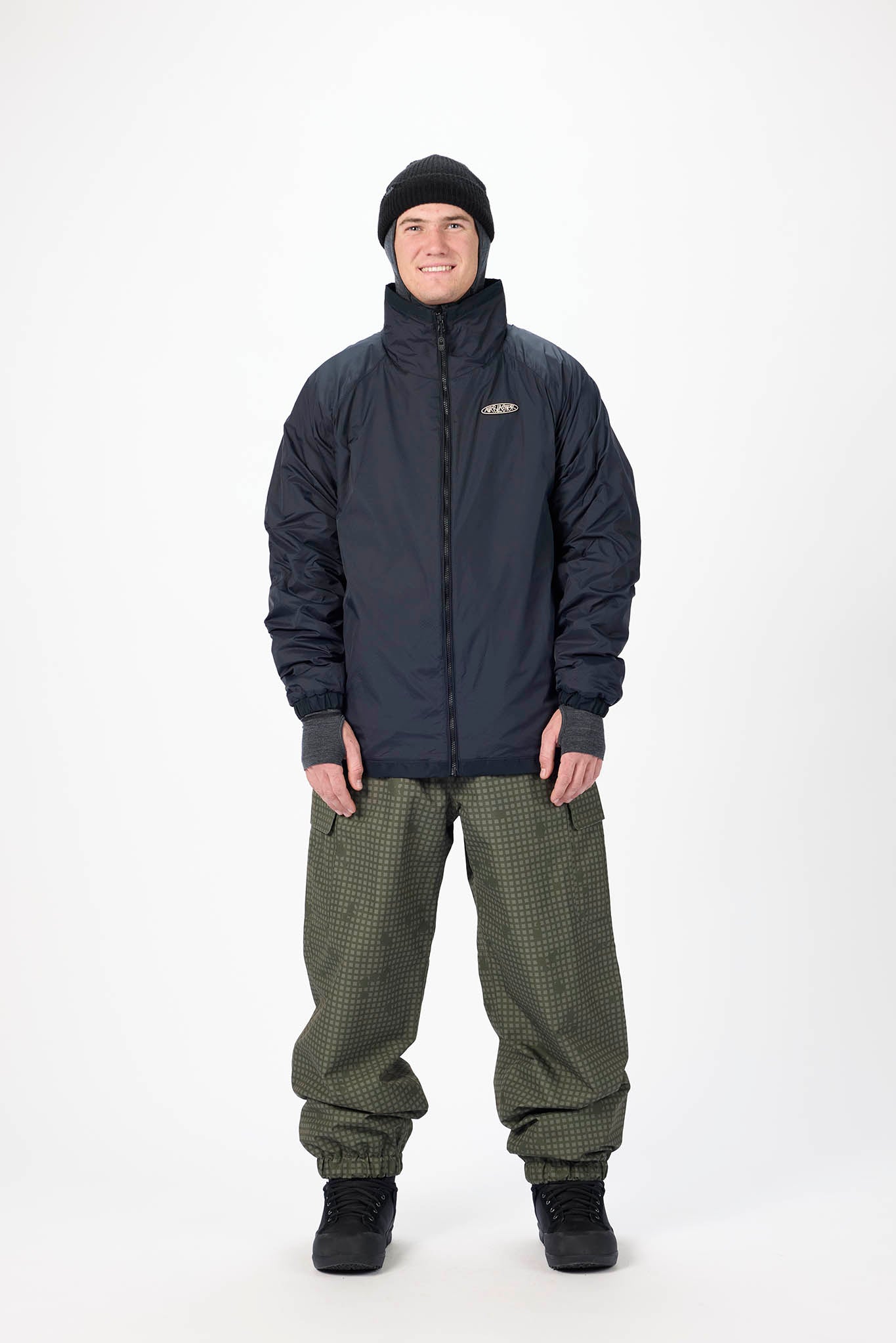 A man wearing a dark navy insulated jacket, olive green patterned snow pants, a black beanie, and black boots, standing against a plain white studio background.