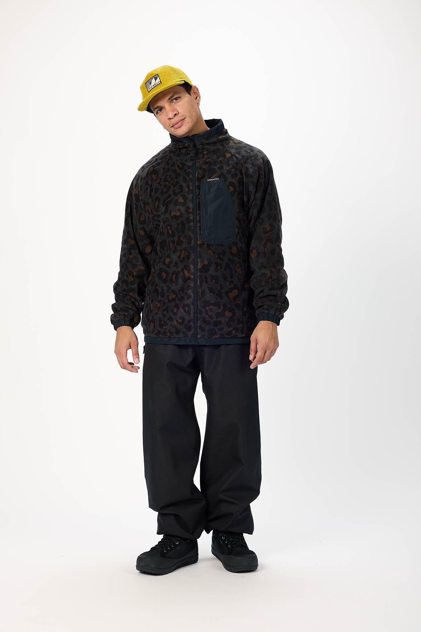 A model wearing a black and dark brown leopard-print fleece jacket with a dark chest pocket, black pants, and a yellow baseball cap, standing against a plain white studio background.