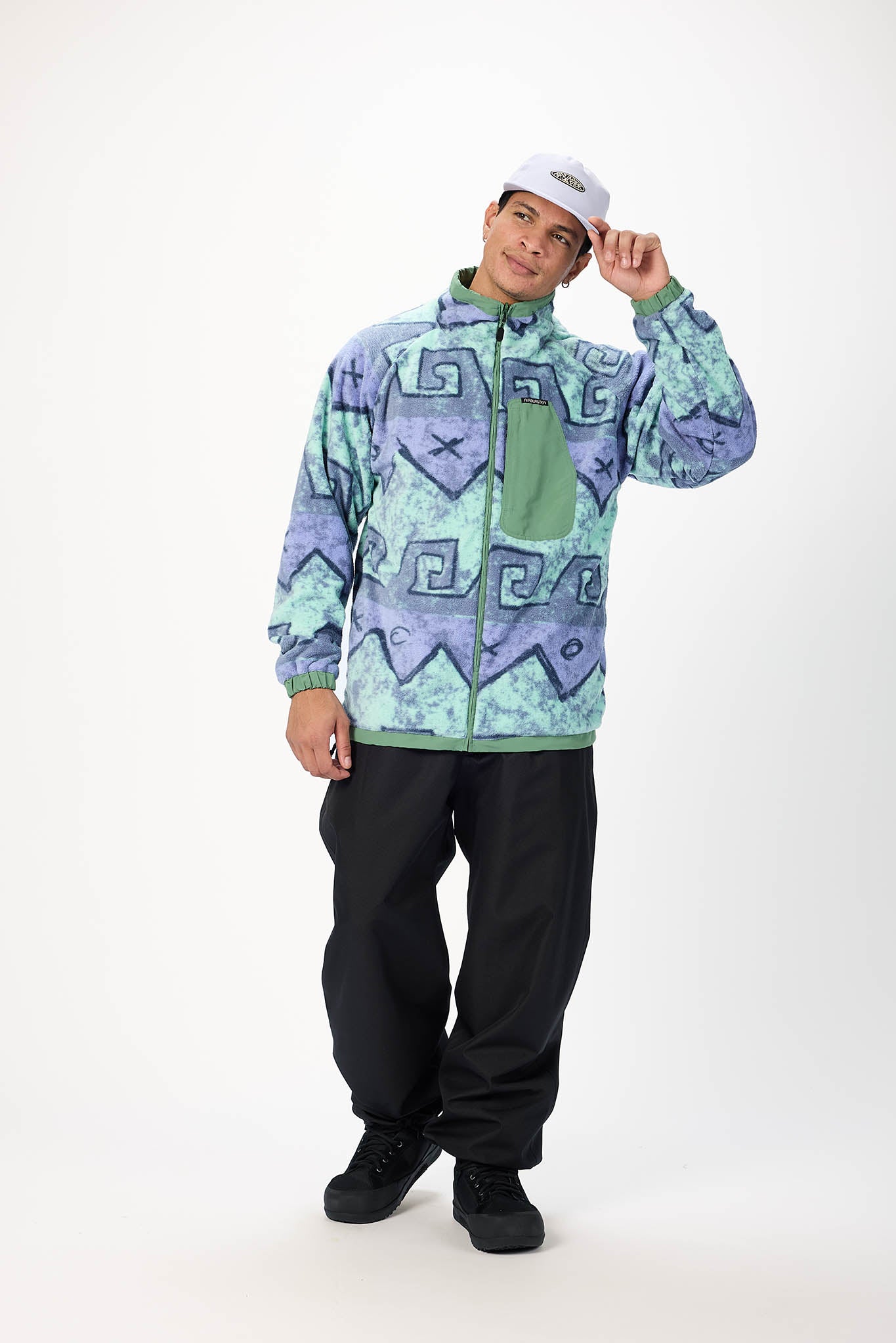 A man modeling a light blue and green patterned zip-up jacket, black snow pants, and a white cap against a plain white studio background.