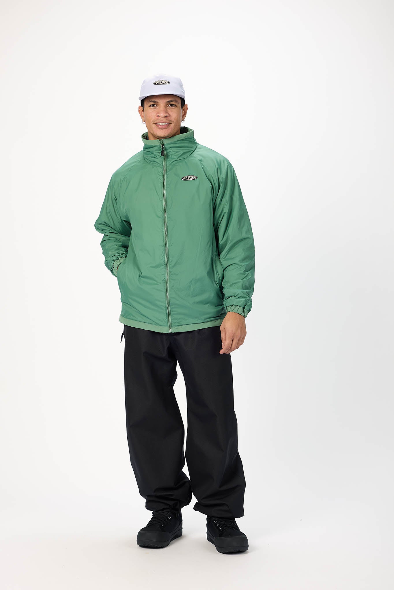 A man wearing a green lightweight zip-up jacket, black pants, black shoes, and a white baseball cap, standing against a plain white studio background.