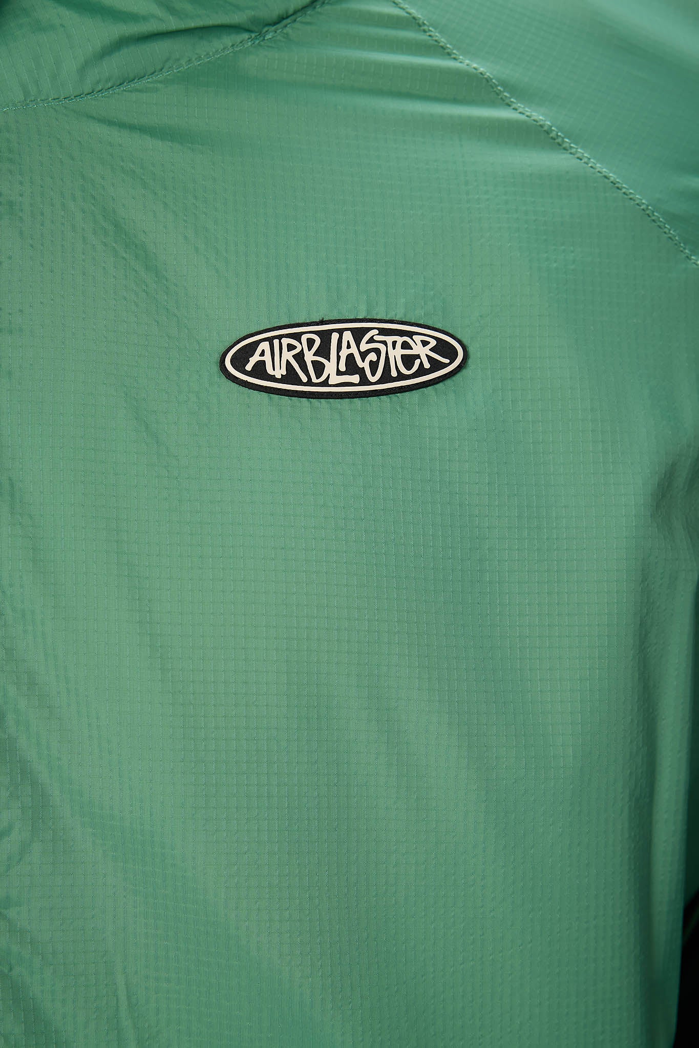 Close-up of a green textured jacket or windbreaker with a black and white Airblaster logo patch on the chest.