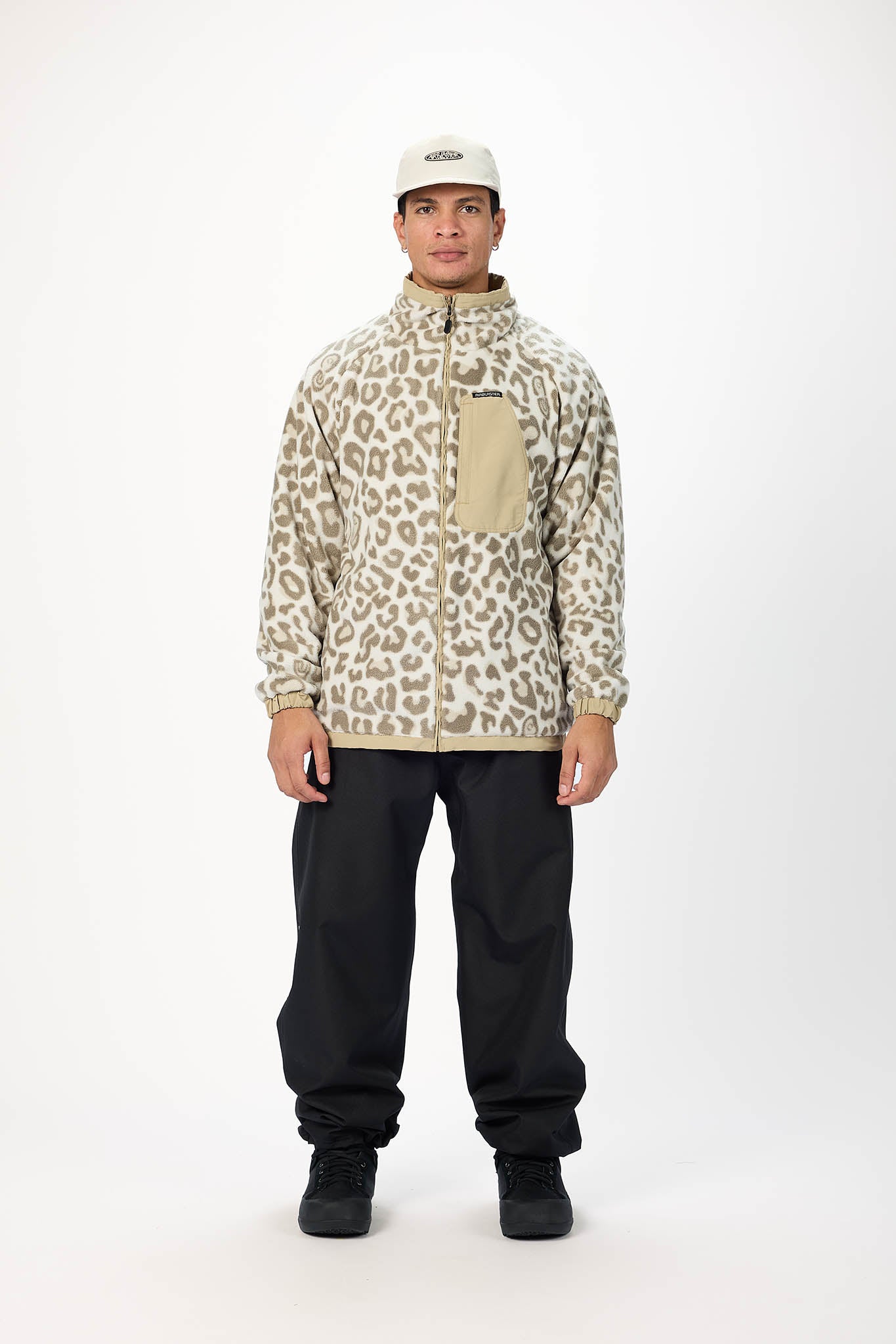 A man modeling a beige and brown leopard-print fleece jacket with a tan chest pocket, paired with black pants and a beige cap, against a plain white studio background.