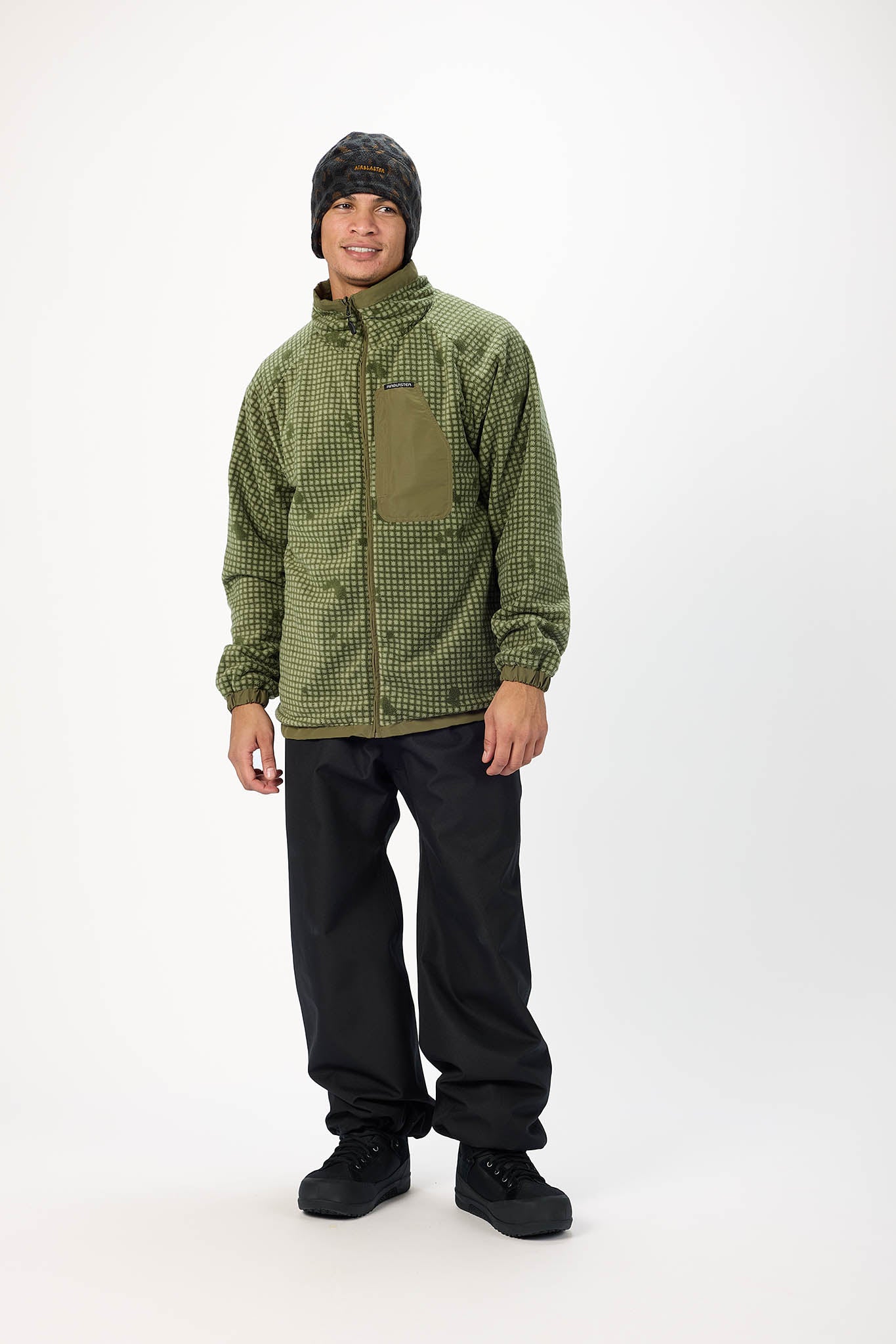 A man wearing an olive green and black checkered fleece jacket with a brown chest pocket, black pants, and a black knit beanie, standing against a white studio background.