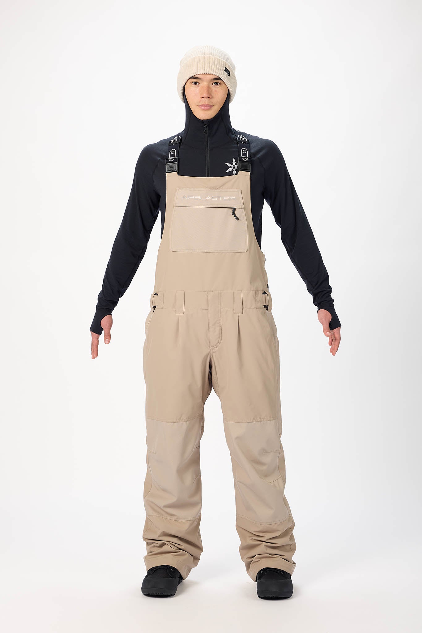 Person wearing beige bib overalls with a black long-sleeve shirt on a white background