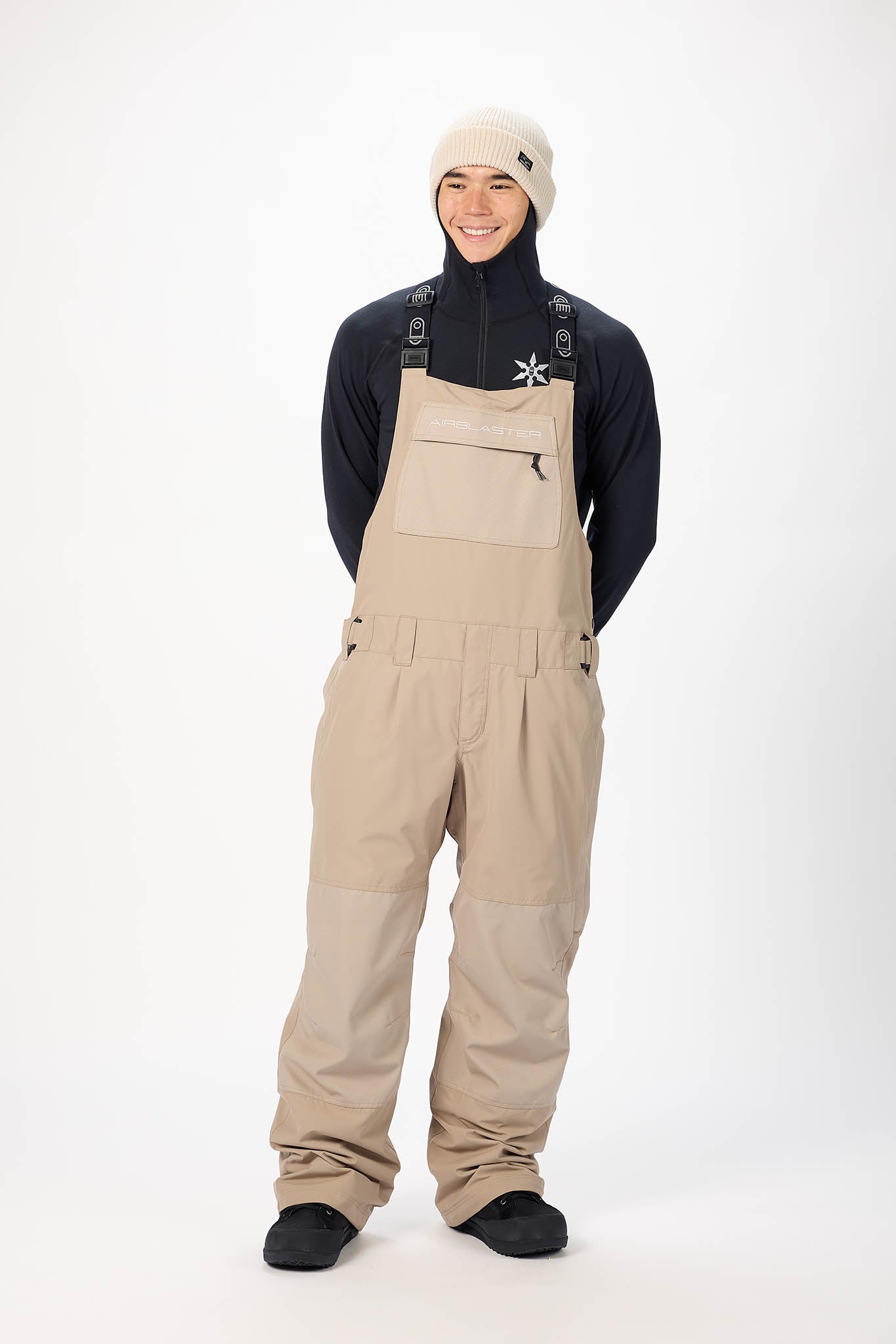 Person wearing beige bib overalls, black shirt, and white beanie on a white background