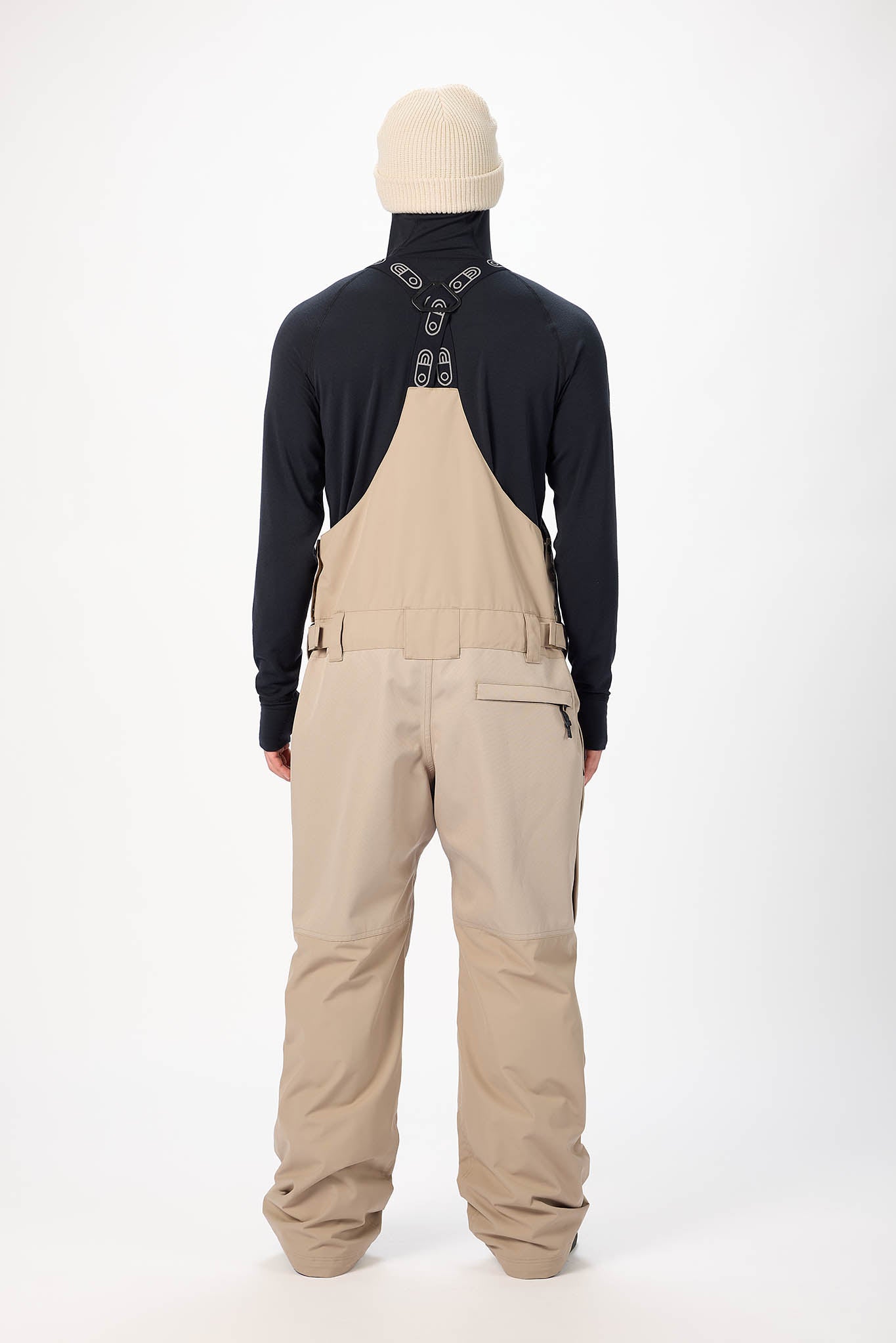 Person wearing beige bib overalls and a black long-sleeve shirt on a white background