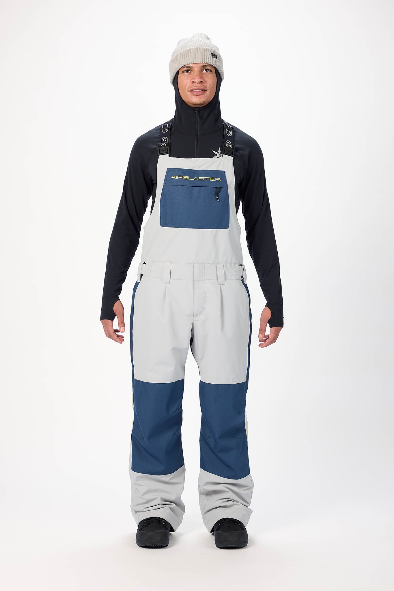 Person wearing a black long-sleeve shirt, white bib overalls with blue accents, and a white knit cap on a white background.