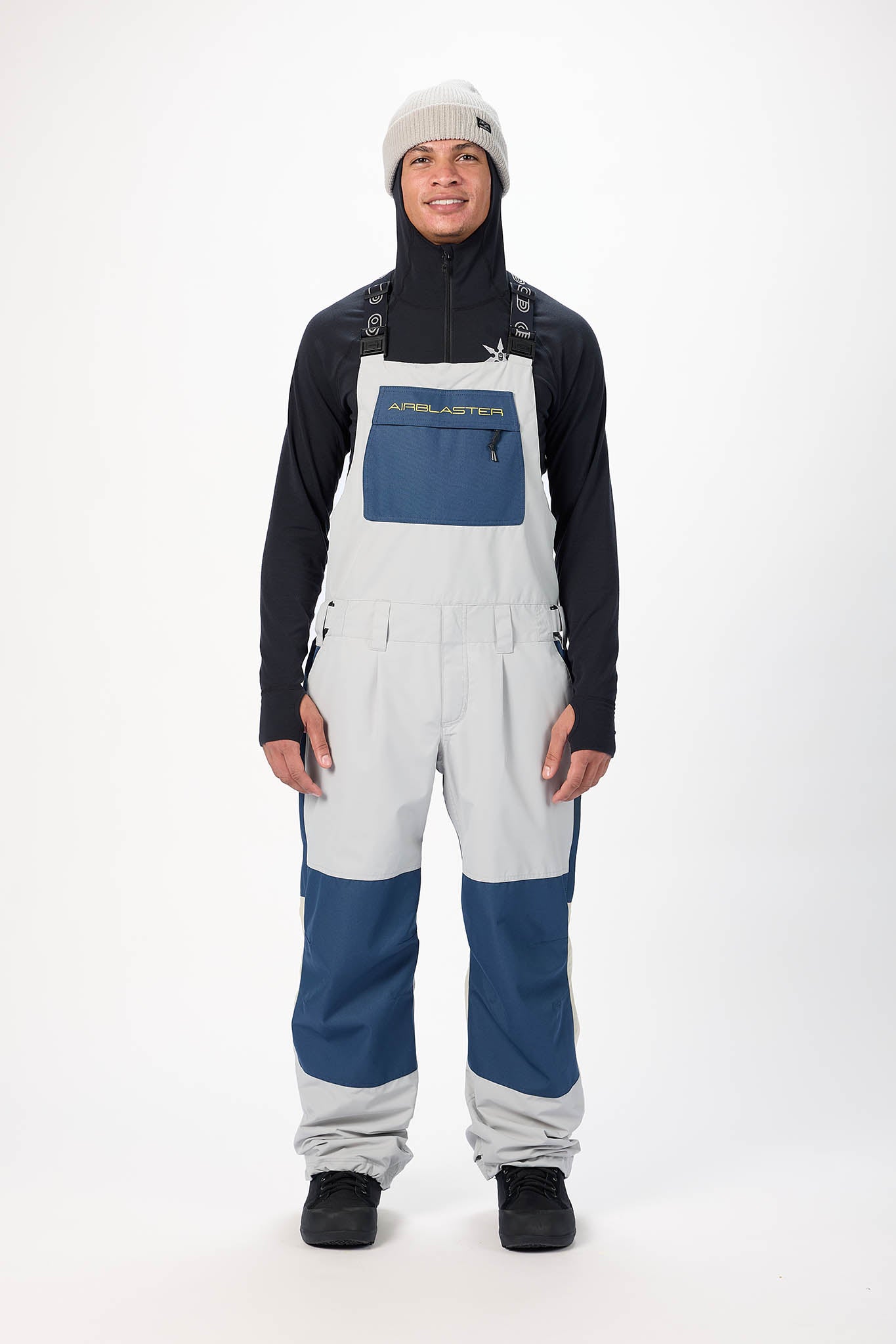 Person wearing a black jacket, white bib overalls with blue accents, and a gray beanie on a white background.
