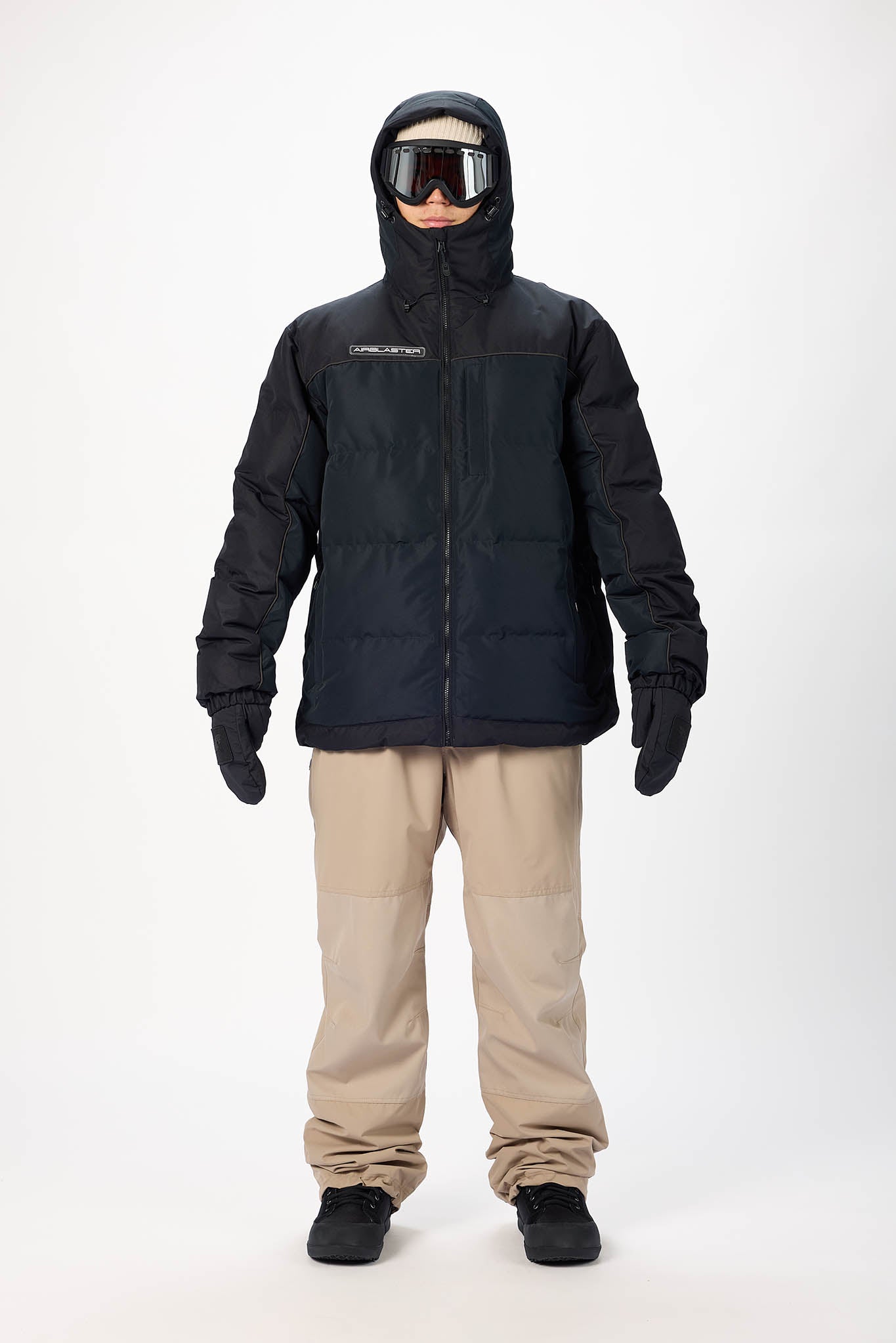 Person wearing a black snow jacket, beige pants, and goggles on a white background