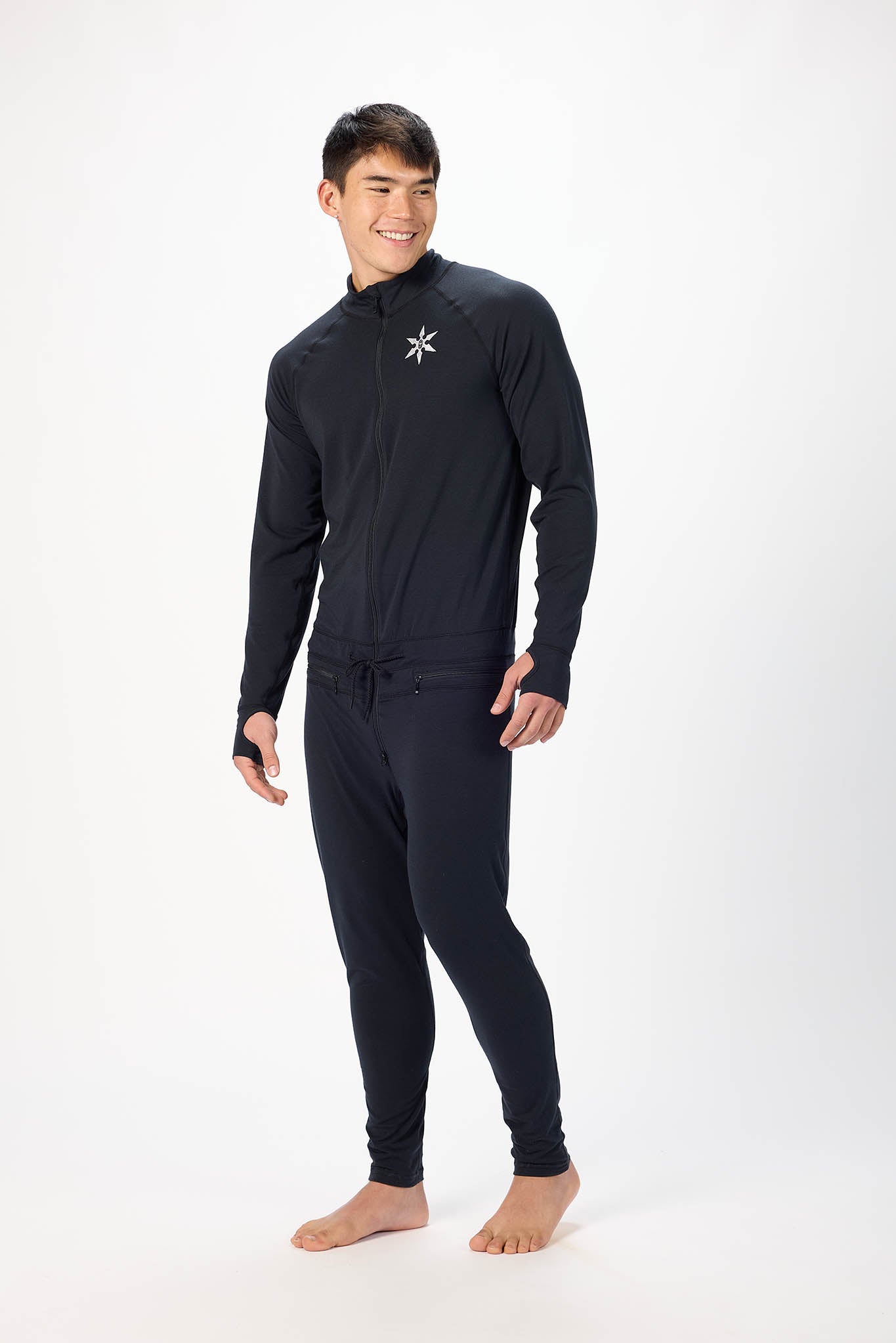 A man modeling a black full-zip athletic onesie with a small white logo on the left chest, standing against a plain white studio background.