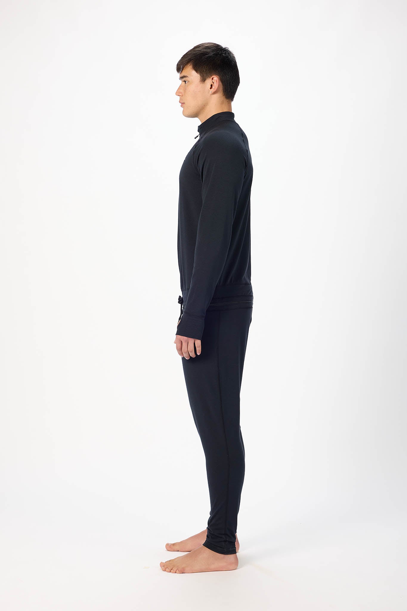 in side profile A man modeling a black full-zip athletic onesie with a small white logo on the left chest, standing against a plain white studio background.