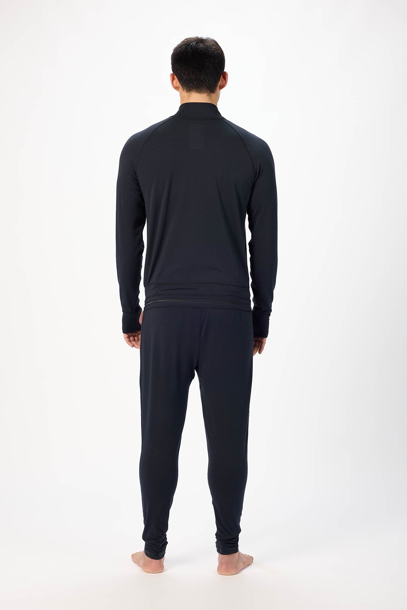 view from behind man modeling a black full-zip athletic onesie with a small white logo on the left chest, standing against a plain white studio background.