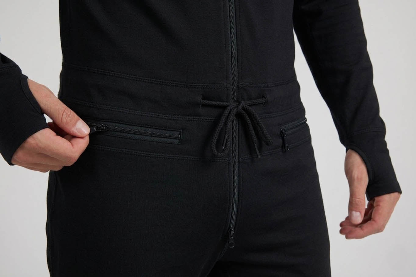 Close-up of a person wearing a black zip-front jacket or romper with a drawstring waist and zip pockets, shown against a light gray studio background.