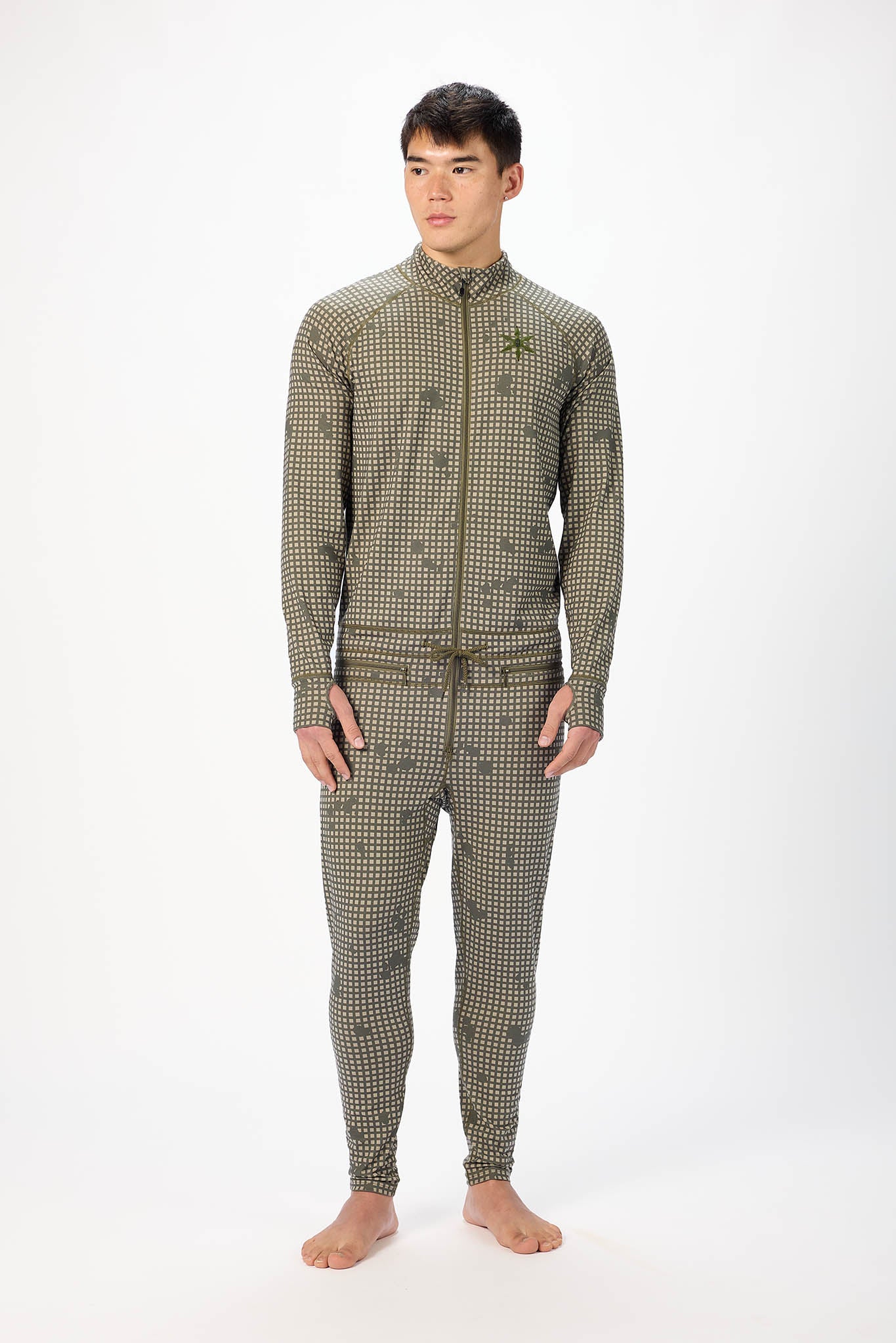 A man modeling a olive grid like pattern full-zip athletic onesie with a small white logo on the left chest, standing against a plain white studio background.