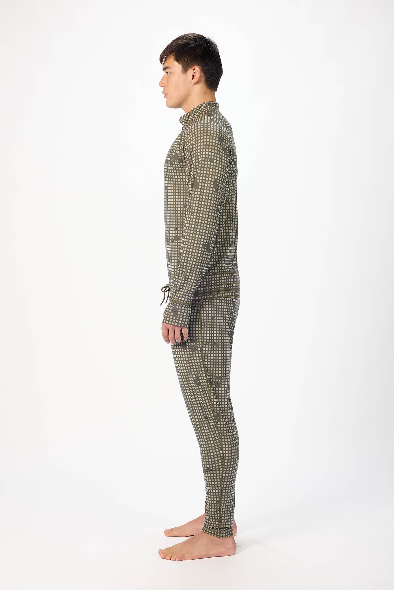 in side profile a man modeling a olive grid like pattern full-zip athletic onesie with a small white logo on the left chest, standing against a plain white studio background.