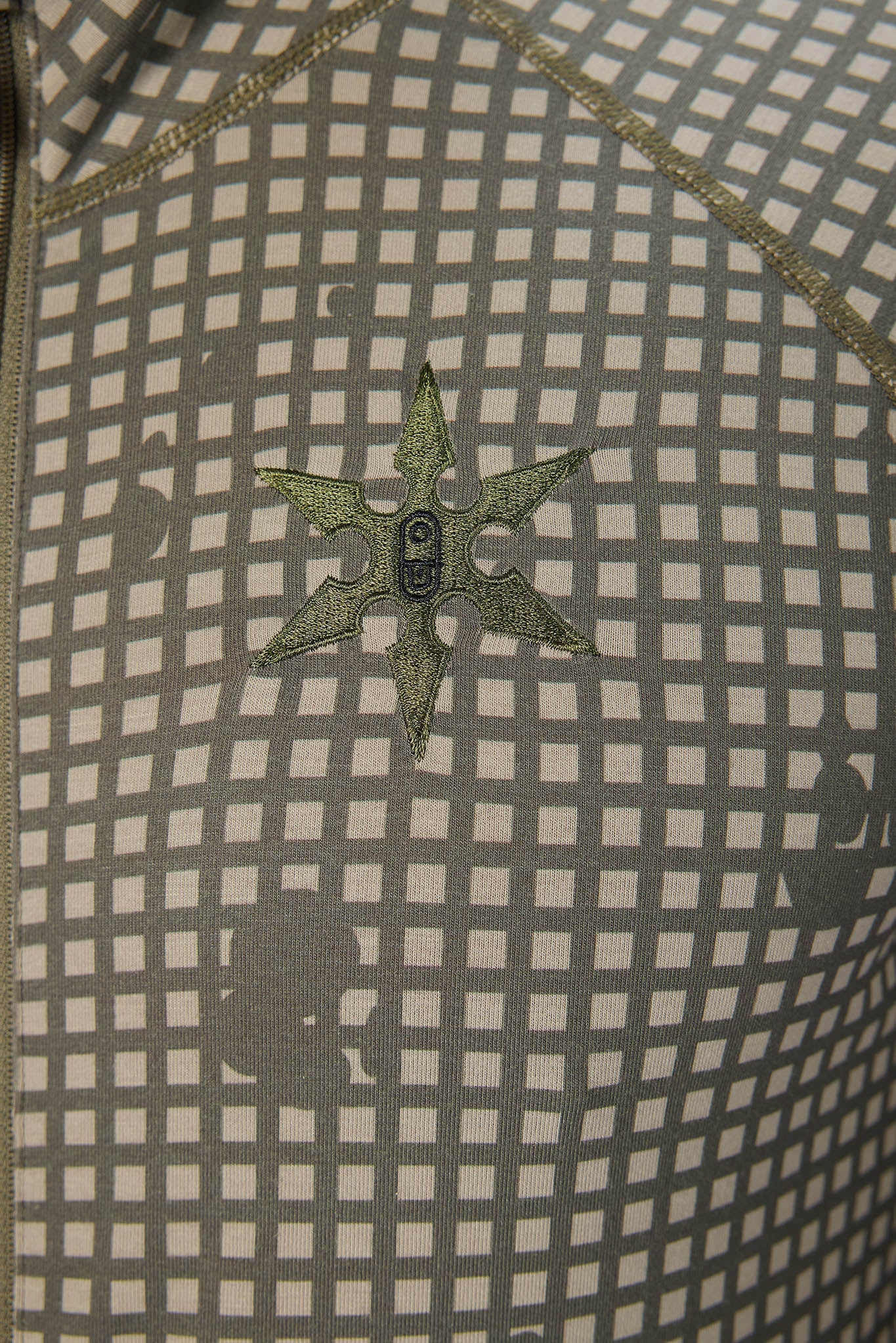 Close-up of a patterned fabric with a gray and beige grid design, featuring a small embroidered star-shaped emblem in olive green in the center.