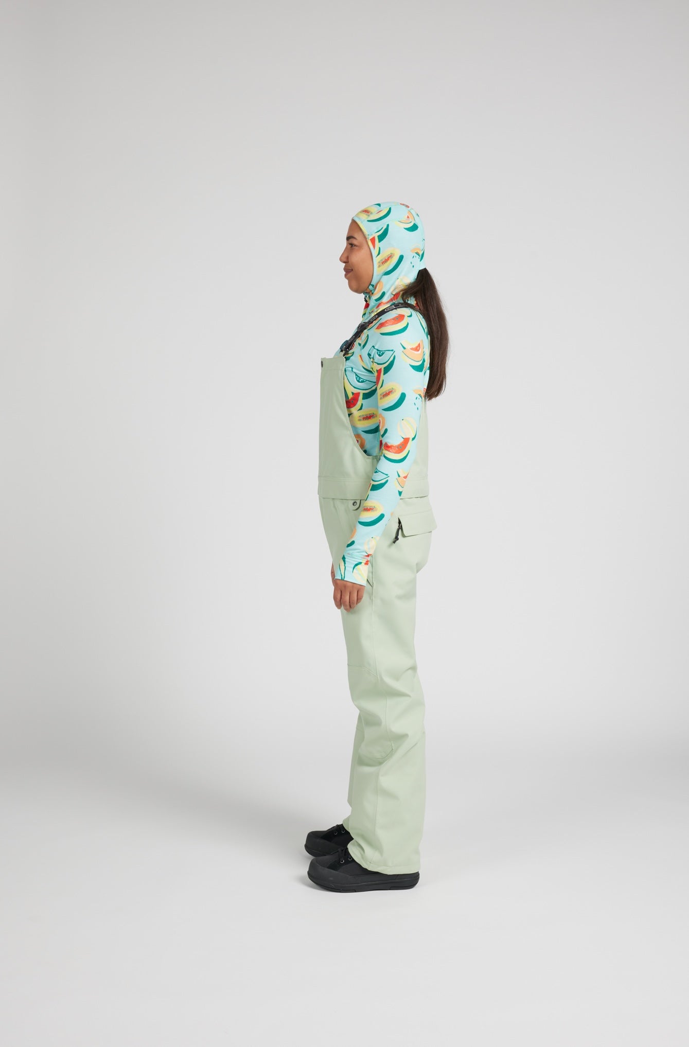 A model shown from the side profile wearing sage green bib overalls with a colorful banana print hood and matching patterned long-sleeve top, paired with black slip-on shoes, against a light gray studio background.