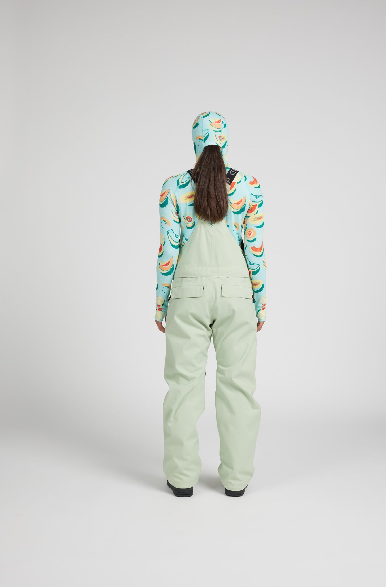 A model shown from the back wearing sage green snow bib overalls with a matching light green hood, paired with a light blue long-sleeve top featuring a colorful tropical print.