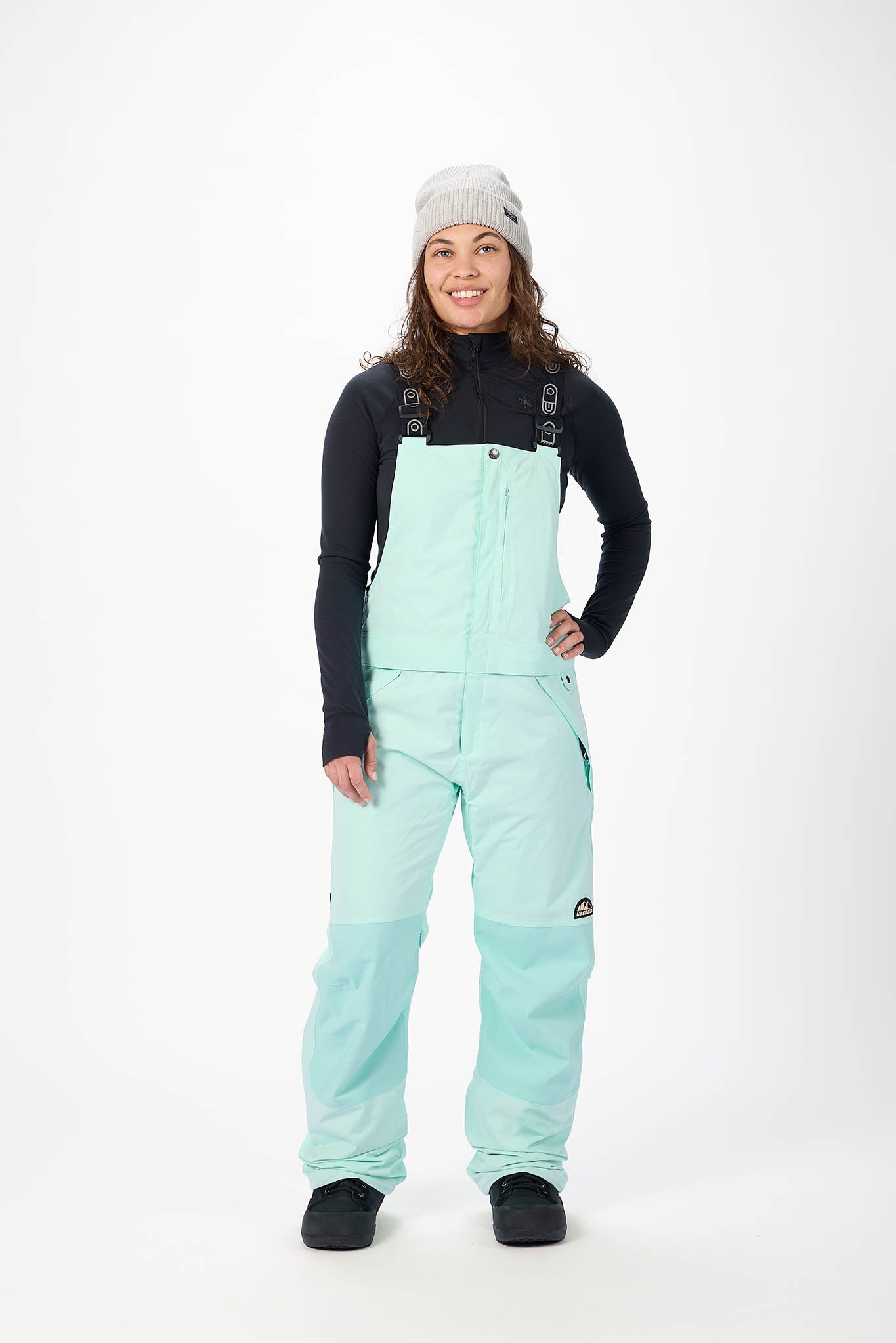 Person wearing a mint green snow bib overalls with a black long-sleeve shirt on a white background