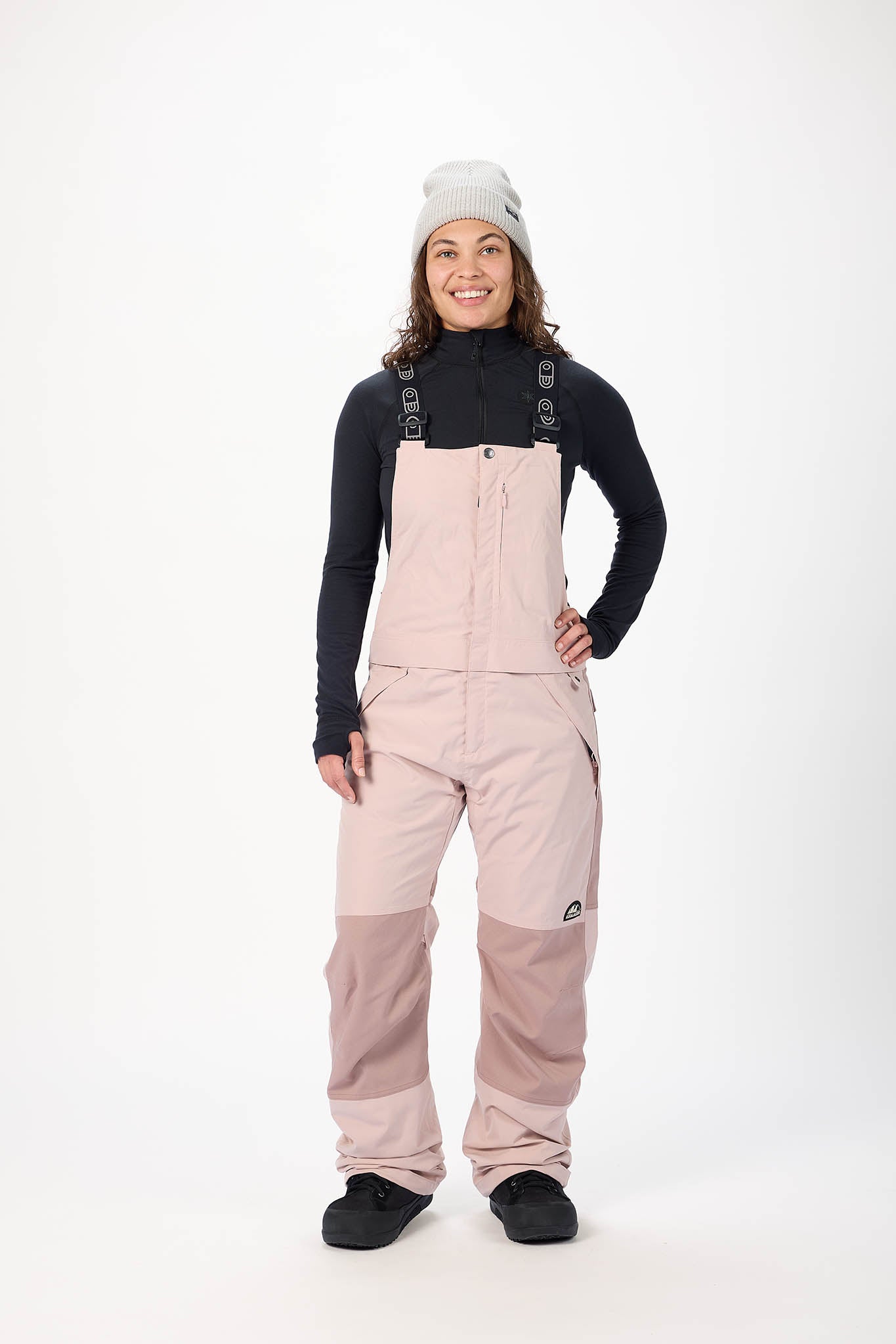 Person wearing a black long-sleeve shirt, beige bib overalls, and black shoes on a white background