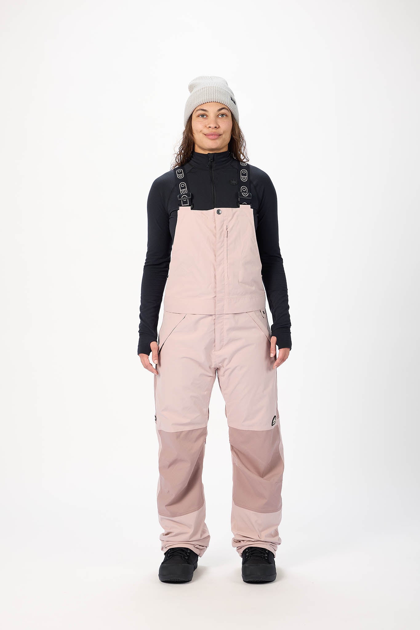 Person wearing a black long-sleeve shirt and pink bib overalls on a white background