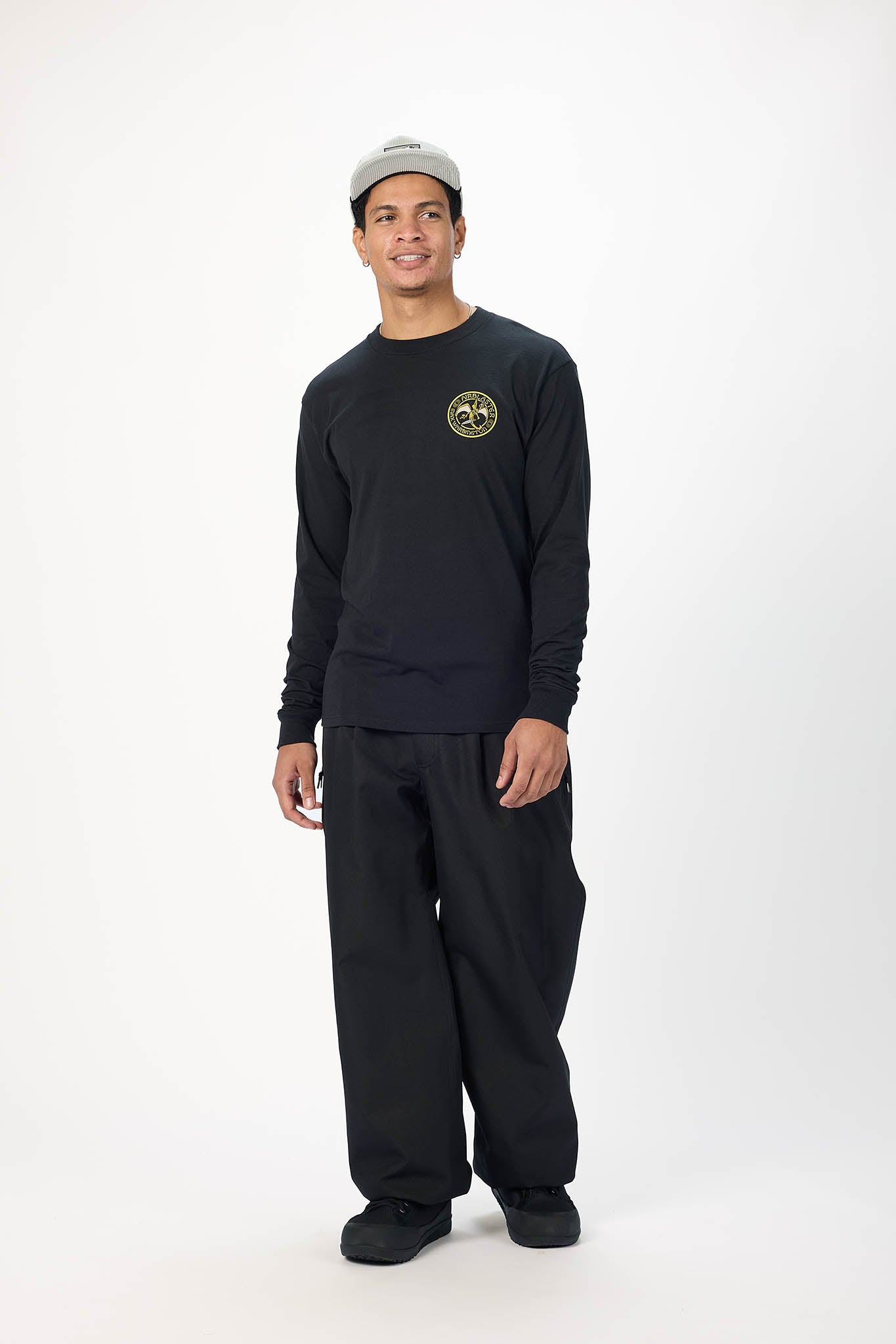 A man modeling a black long-sleeve crewneck sweatshirt with a circular gold logo on the left chest, paired with black sweatpants and a light gray baseball cap, against a plain white studio background.