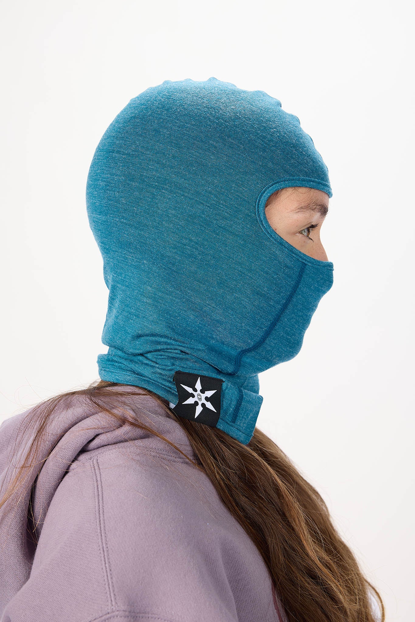 A side-profile view of a person wearing a teal blue heathered balaclava hood with a black and white logo patch on the neck, paired with a lavender hoodie against a plain white background.