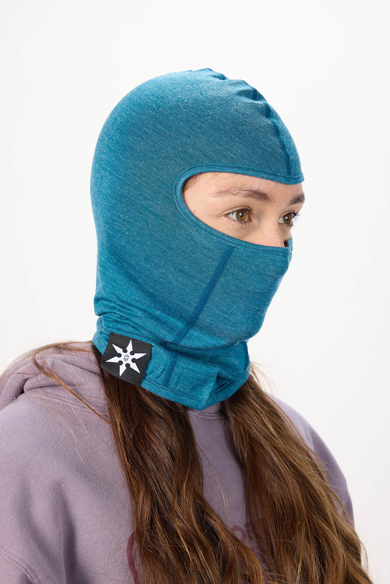 A person wearing a teal blue heathered balaclava hood with a black and white logo patch on the neck, paired with a lavender hoodie against a plain white background.