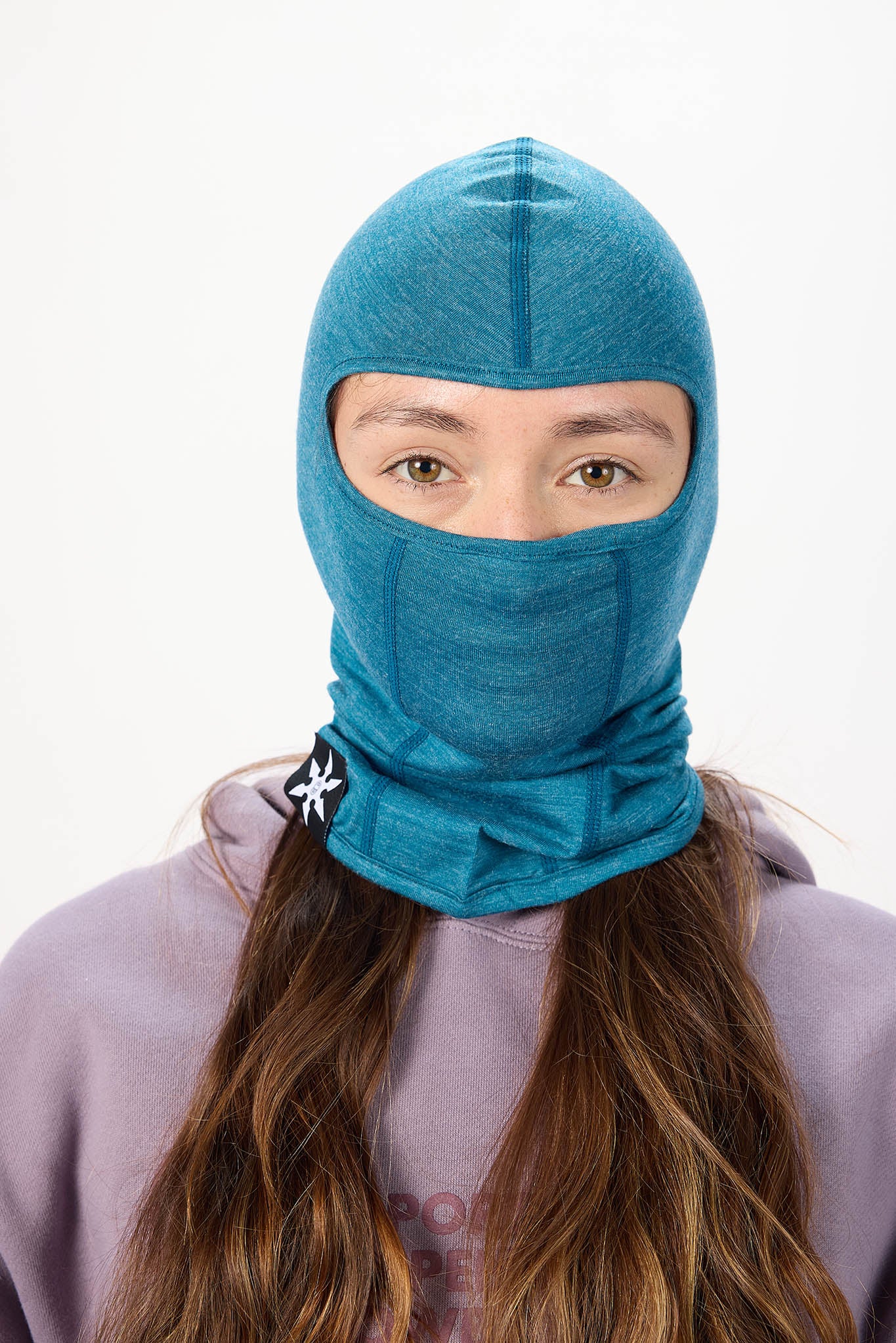 A woman wearing a teal blue heathered balaclava hood and neck gaiter, paired with a lavender hoodie, against a plain white studio background.