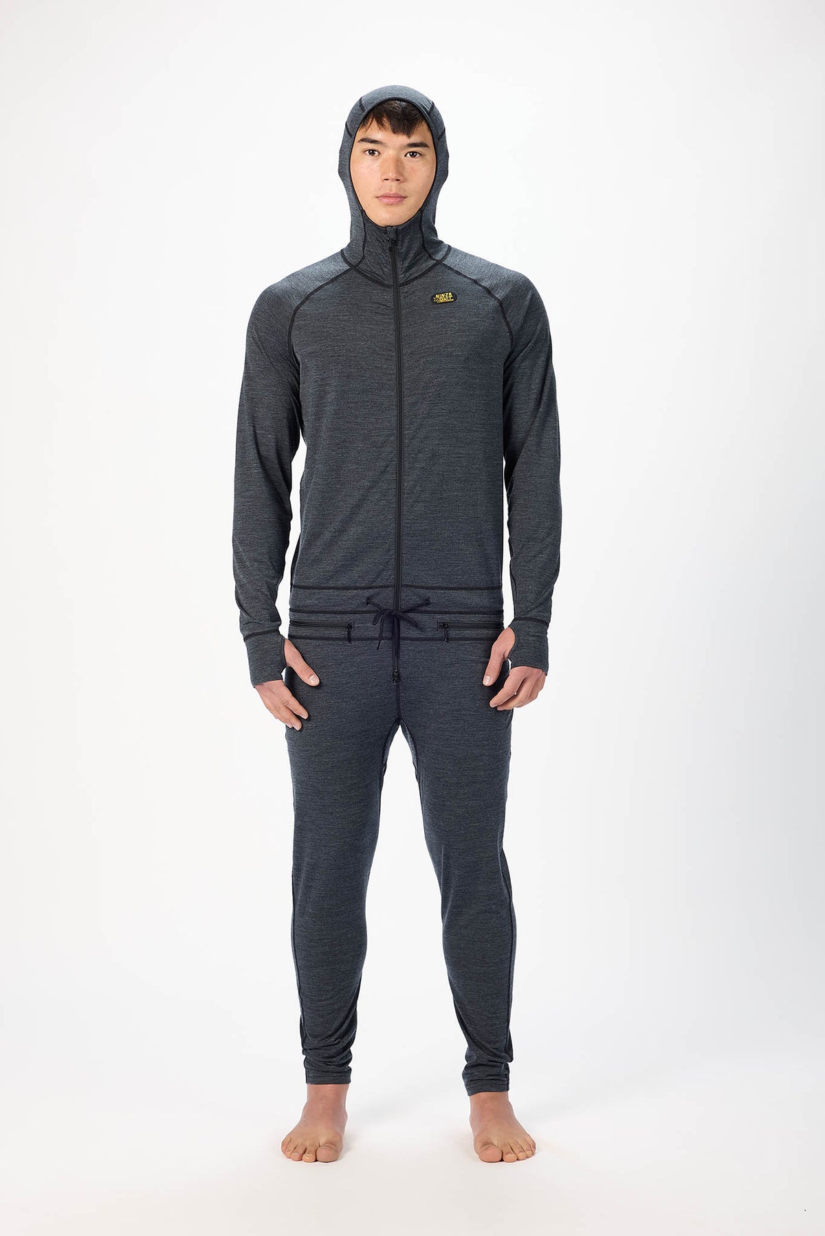 A man wearing a black hooded zip-up onesie , standing barefoot against a plain white studio background.