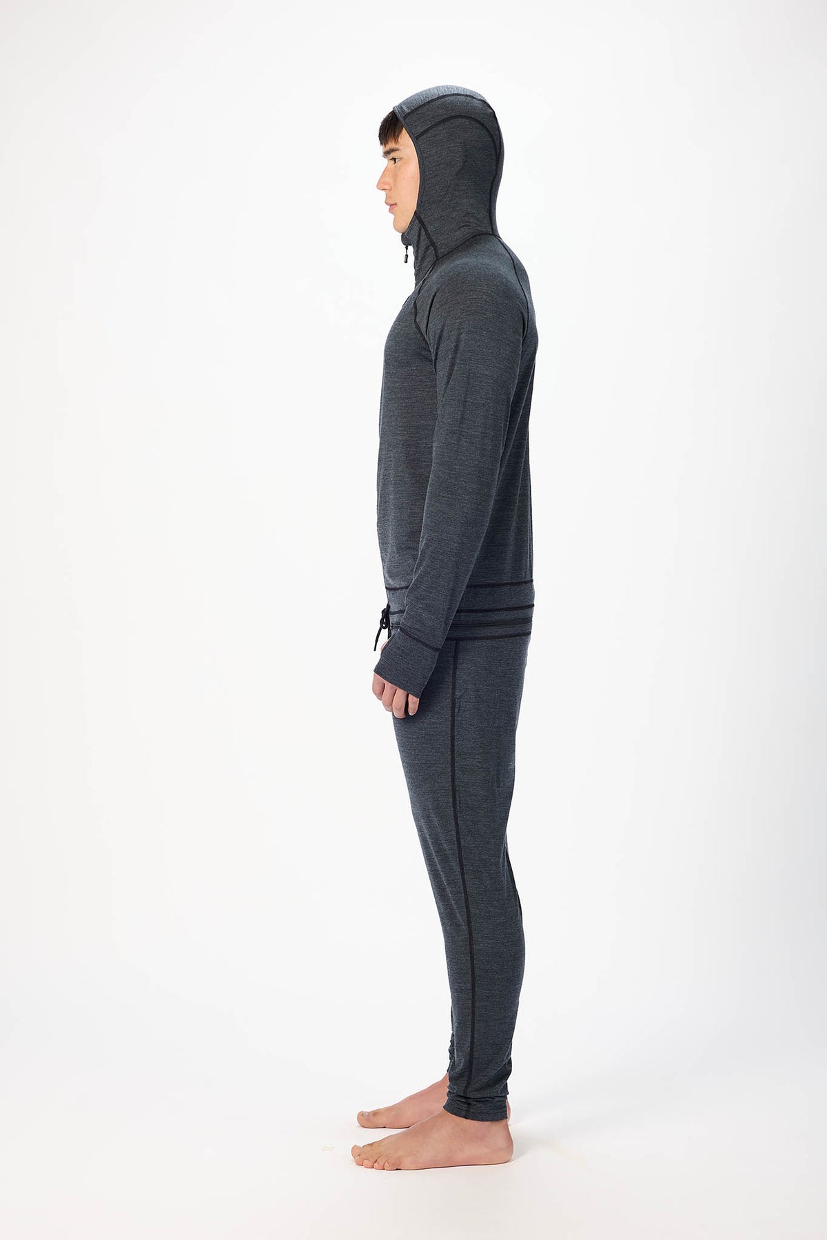 in side profile A man wearing a black hooded zip-up onesie , standing barefoot against a plain white studio background.