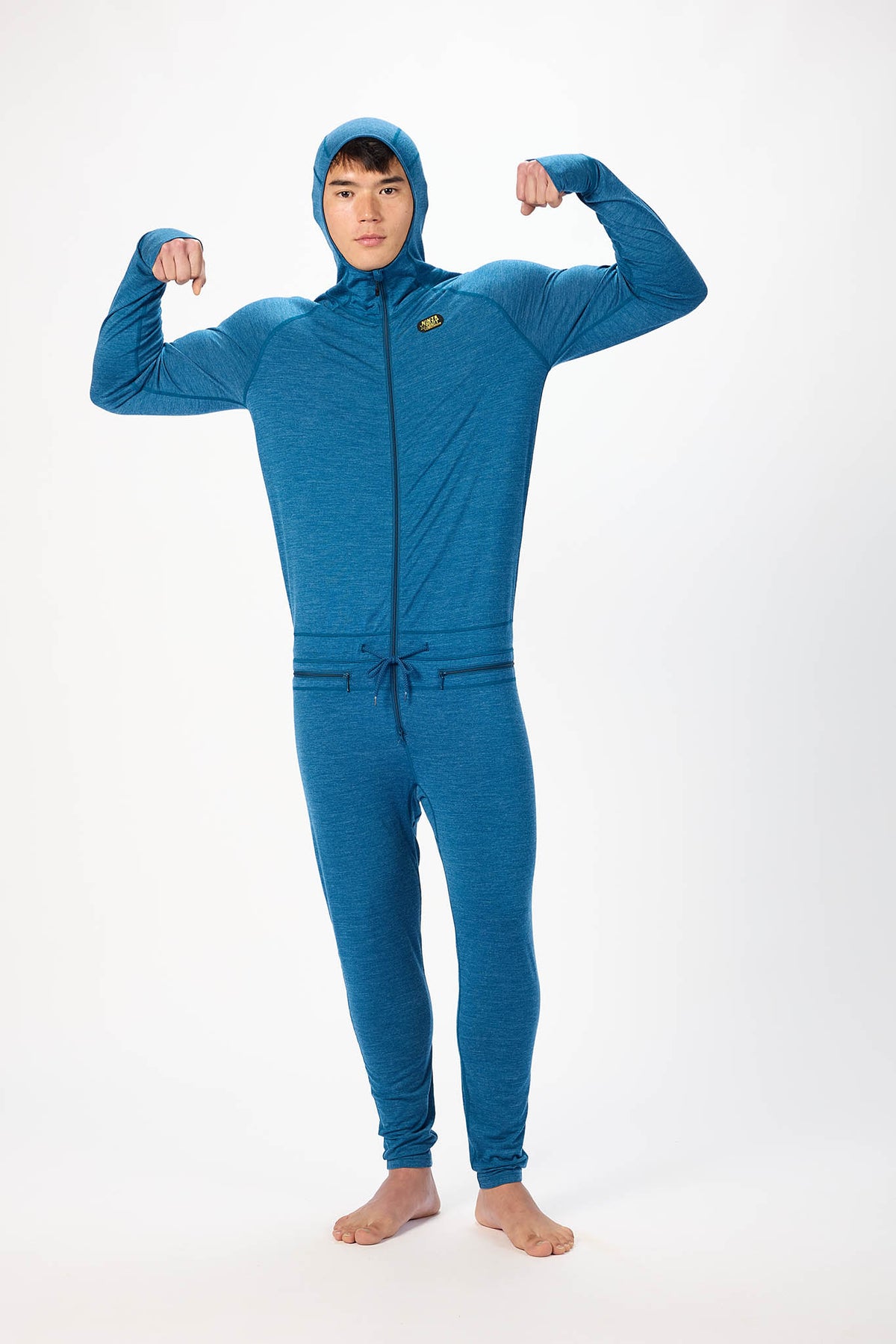 A man wearing a dark gray hooded zip-up onesie , standing barefoot against a plain white studio background. flexing