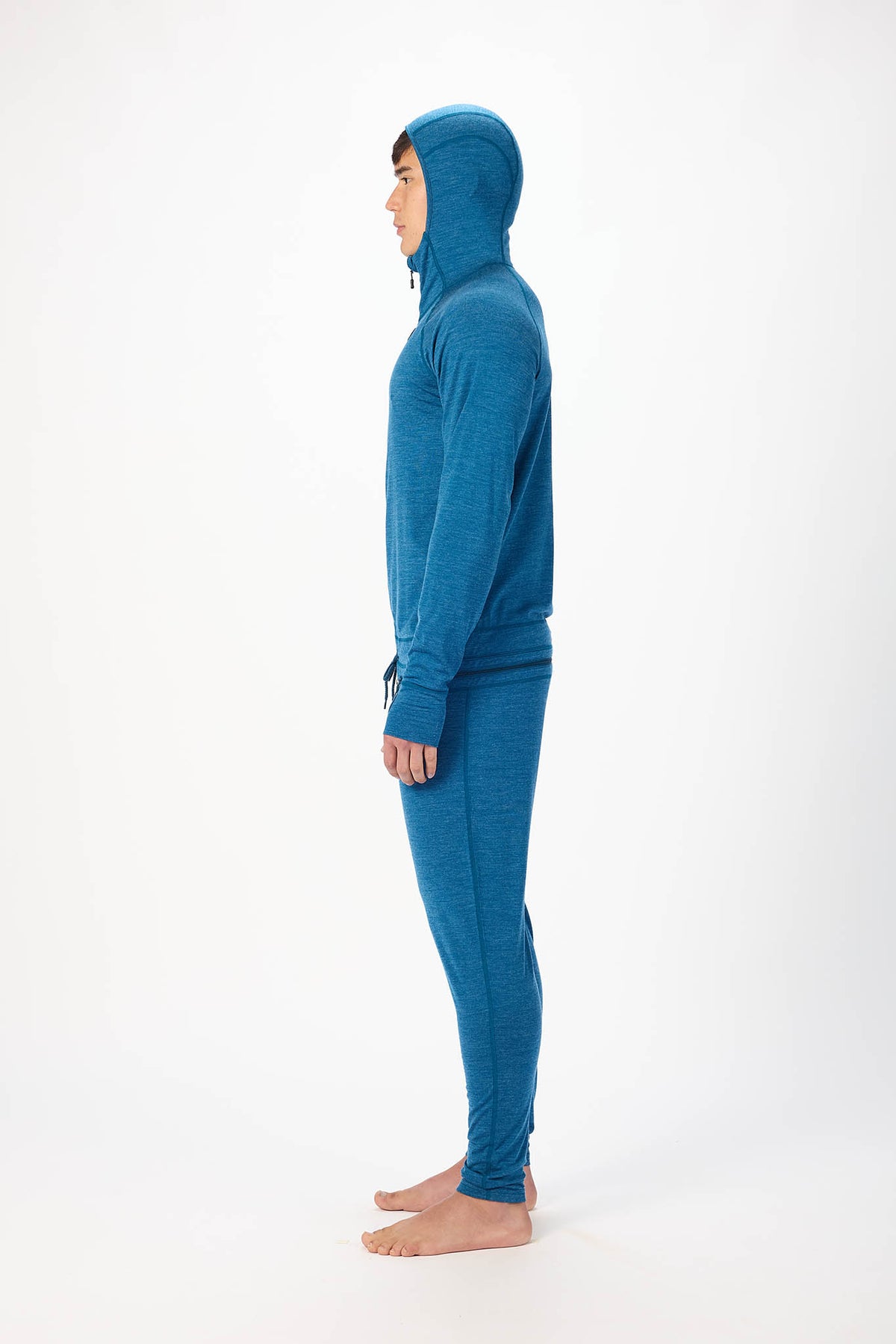 A man wearing a blue hooded zip-up onesie , standing barefoot against a plain white studio background.