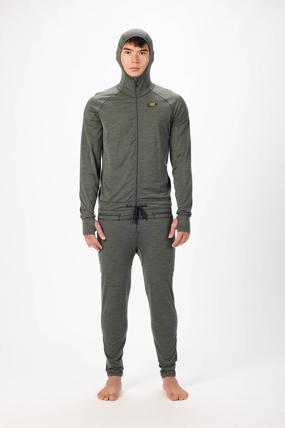 A man wearing a dark gray hooded zip-up onesie , standing barefoot against a plain white studio background.