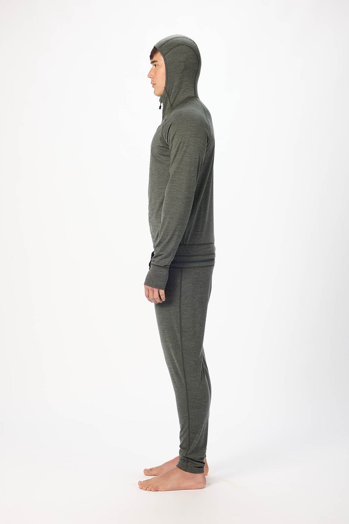 in side profile A man wearing a dark gray hooded zip-up onesie , standing barefoot against a plain white studio background.