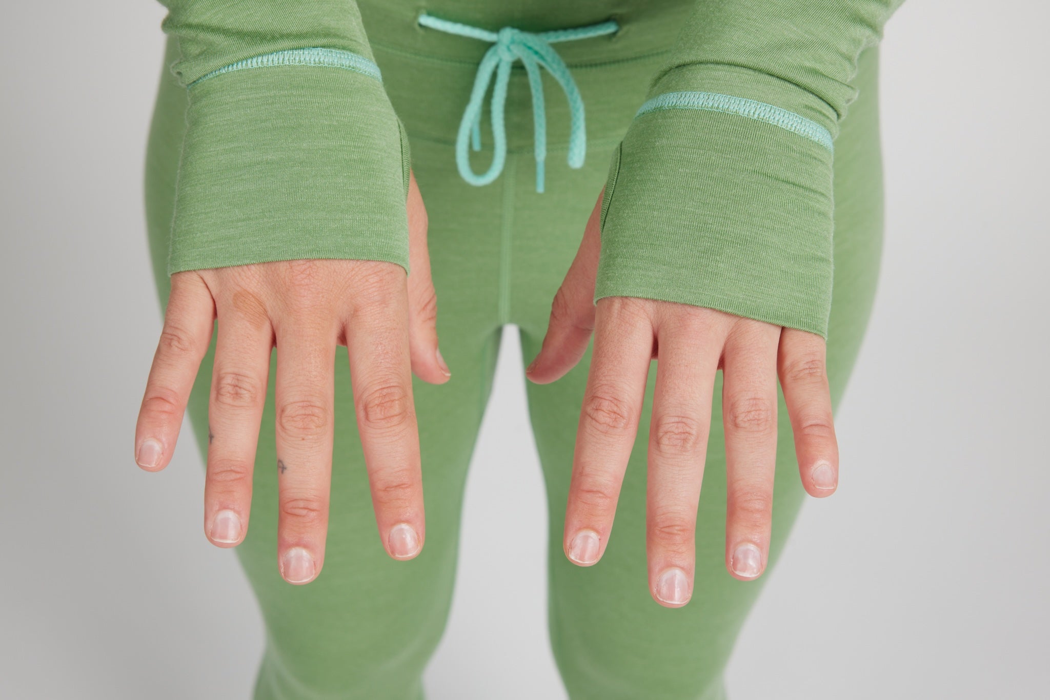 A close-up of a person wearing a sage green ribbed knit lounge set, featuring fingerless cuffs on the long-sleeve top and matching pants, with a mint green drawstring at the waist. The person's hands are shown against a neutral light gray background.